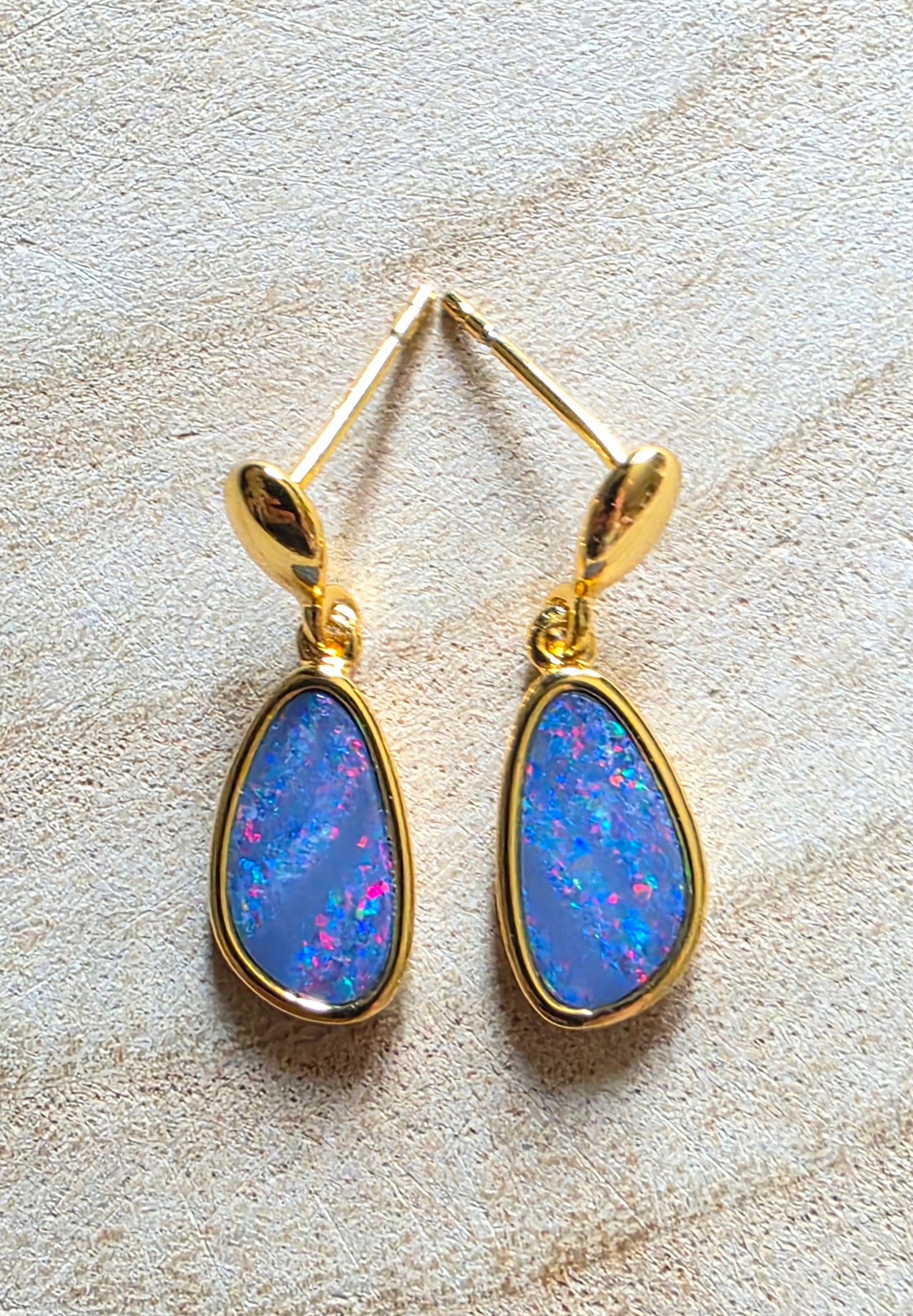 A pair of gold drop earrings with teardrop-shaped blue opal gemstones, displayed on a textured beige surface.