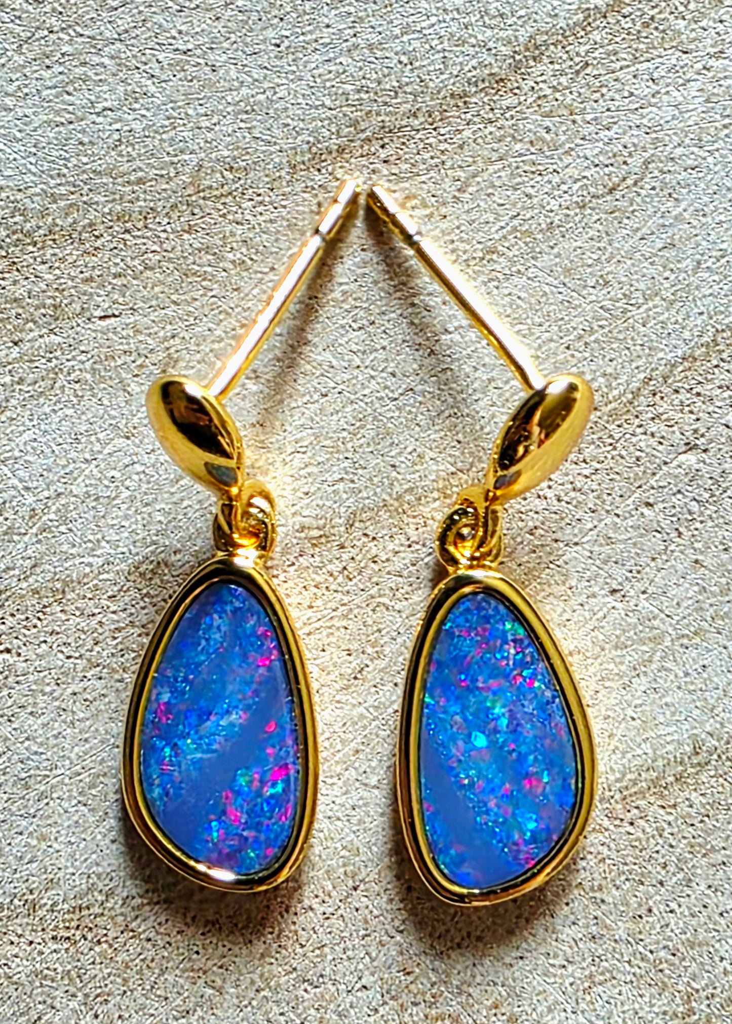 A pair of gold drop earrings with teardrop-shaped blue opal gemstones, displayed on a textured beige surface.