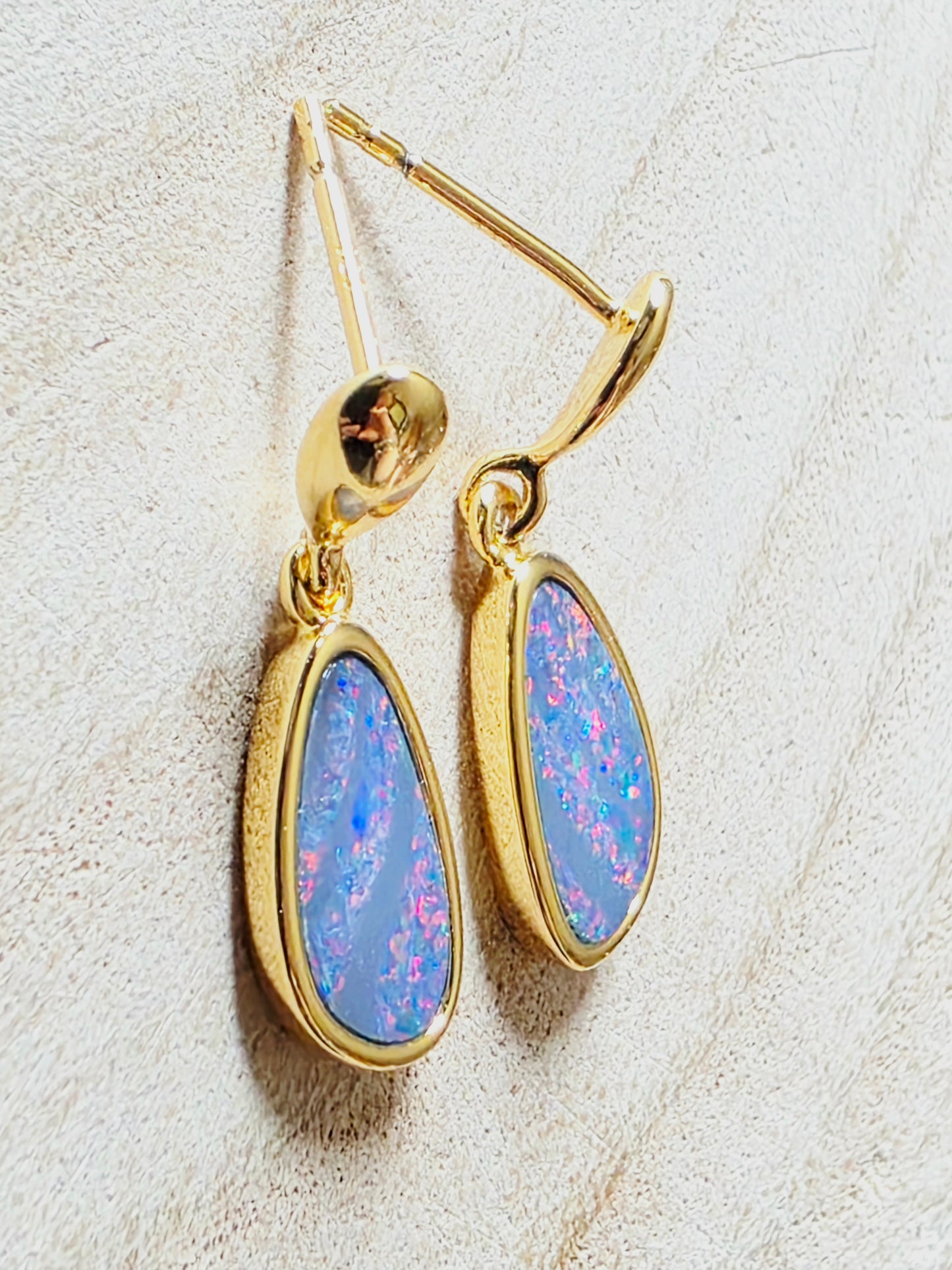 A pair of gold drop earrings with teardrop-shaped blue opal gemstones, displayed on a textured off-white surface.