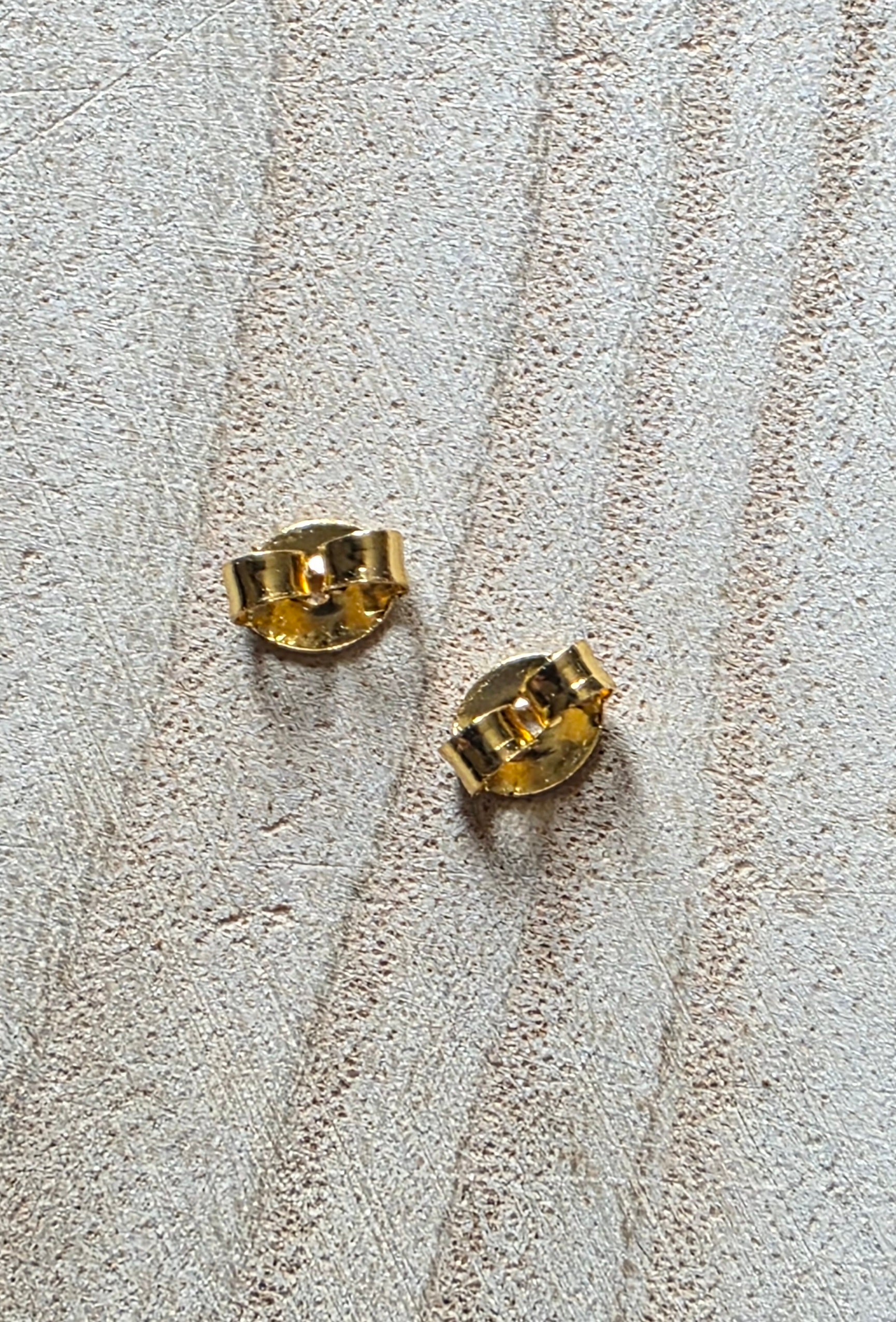 A pair of small gold stud earrings shown from the back on a textured beige fabric background.