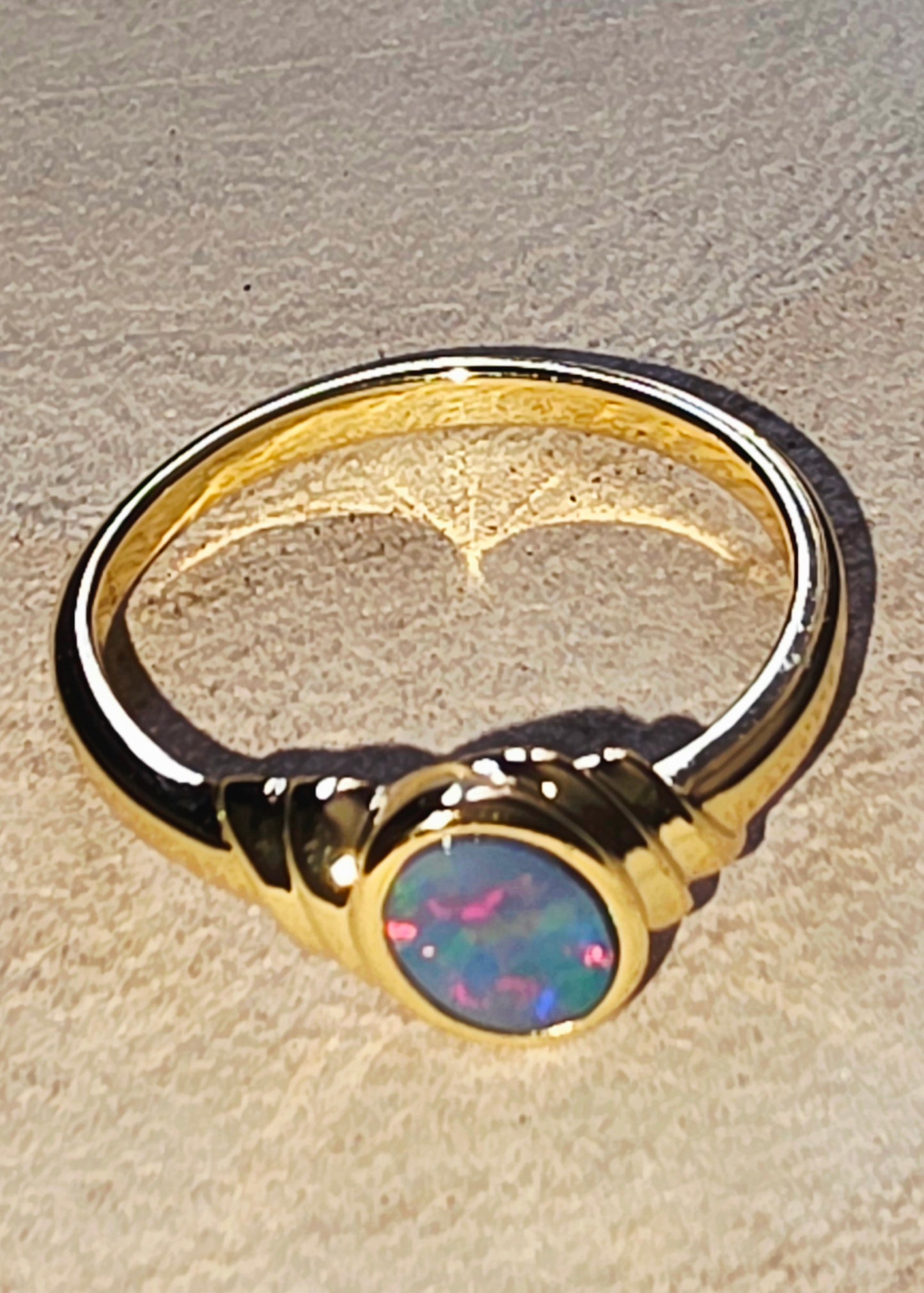 A gold ring with a round iridescent opal gemstone, resting on a textured beige surface.