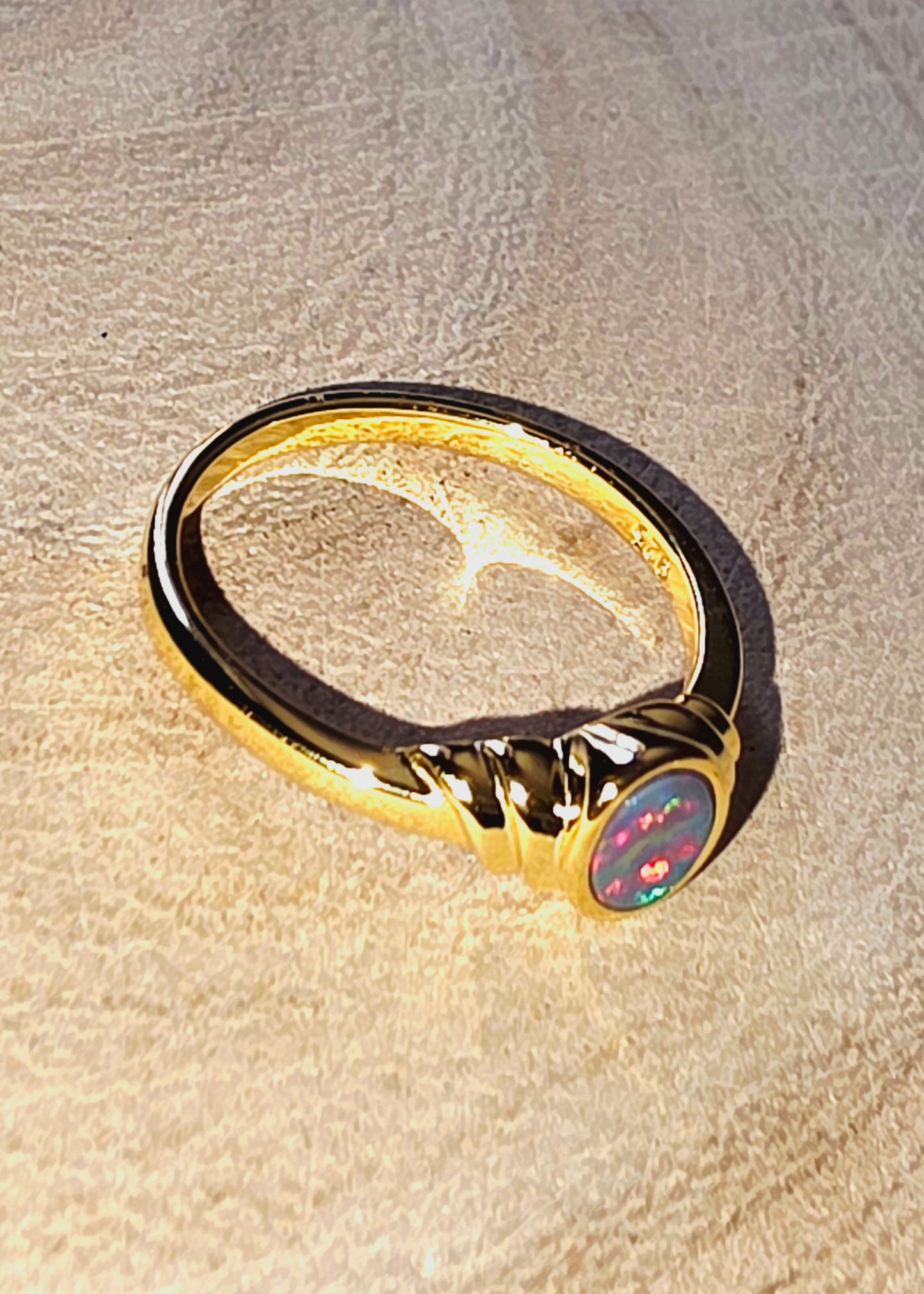 A gold ring with a twisted band and a round cabochon opal featuring a red,  pink, purple, and blue iridescent pattern, resting on a textured beige surface.