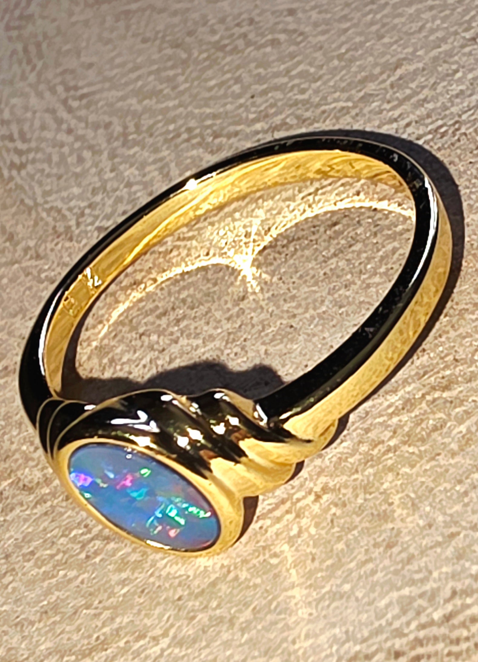 A gold ring featuring an oval cabochon opal with vibrant blue, green, and pink flashes, set against a textured beige background.