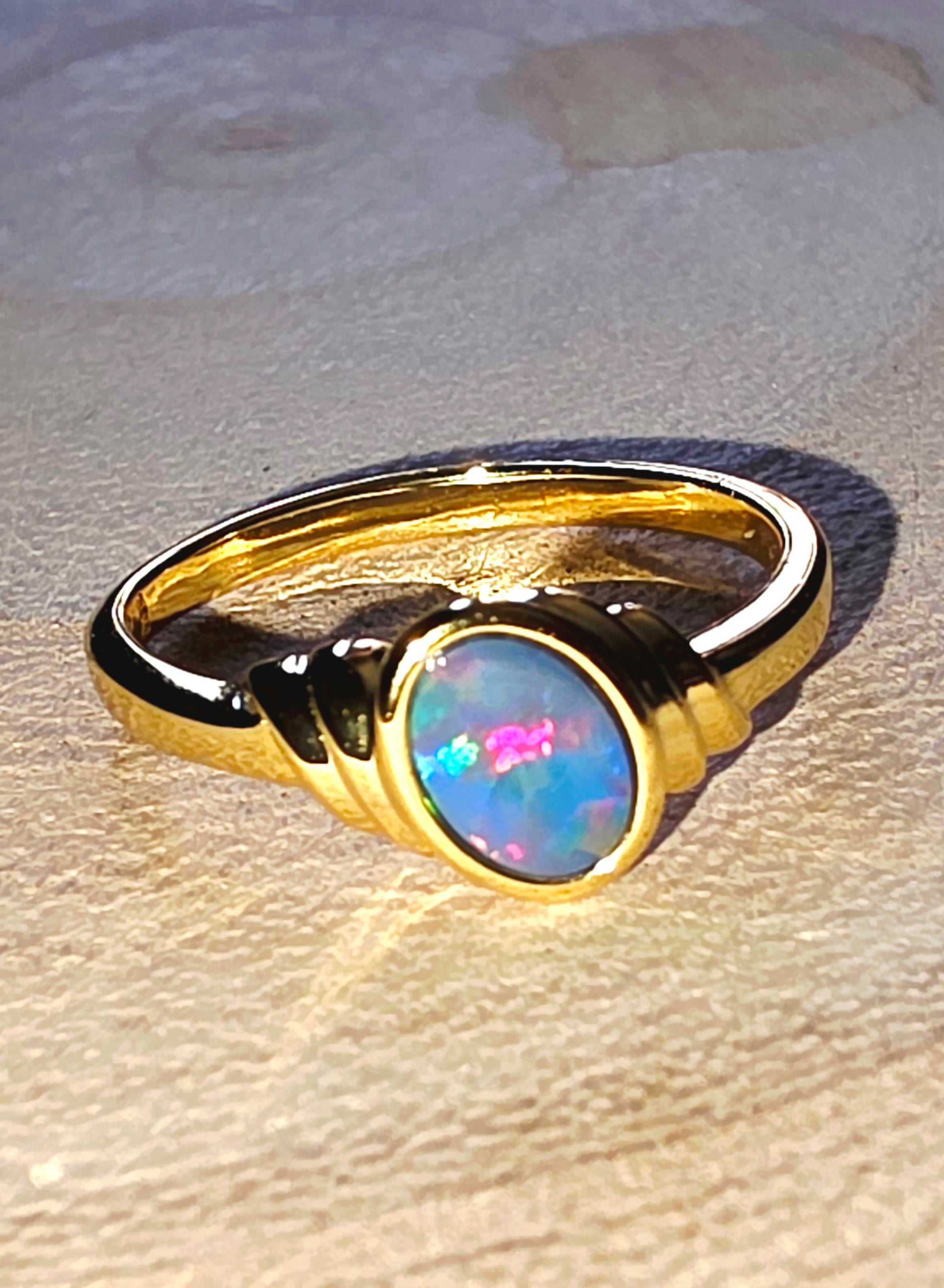 A gold ring featuring a round cabochon opal gemstone with vibrant blue, pink, and green flashes, set against a textured beige background.
