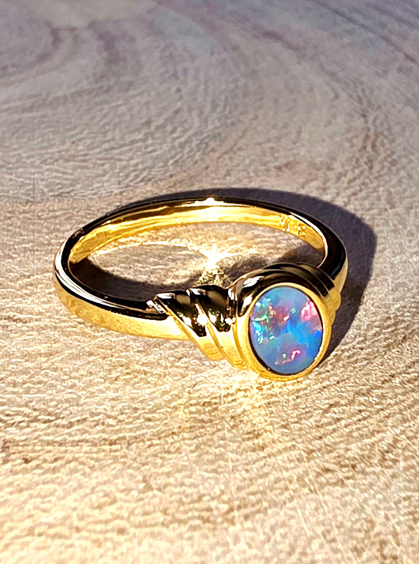 A gold ring with a round iridescent opal gemstone, resting on a textured beige surface under direct sunlight.