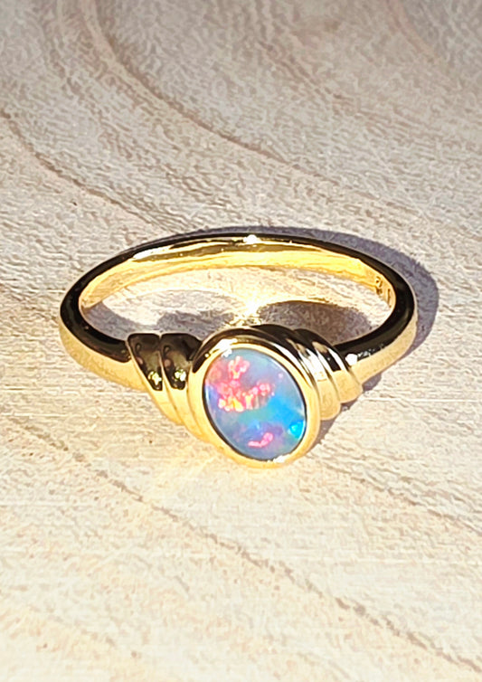 A gold ring featuring an oval cabochon opal gemstone with vibrant blue, pink, and green flashes, displayed on a textured beige surface.