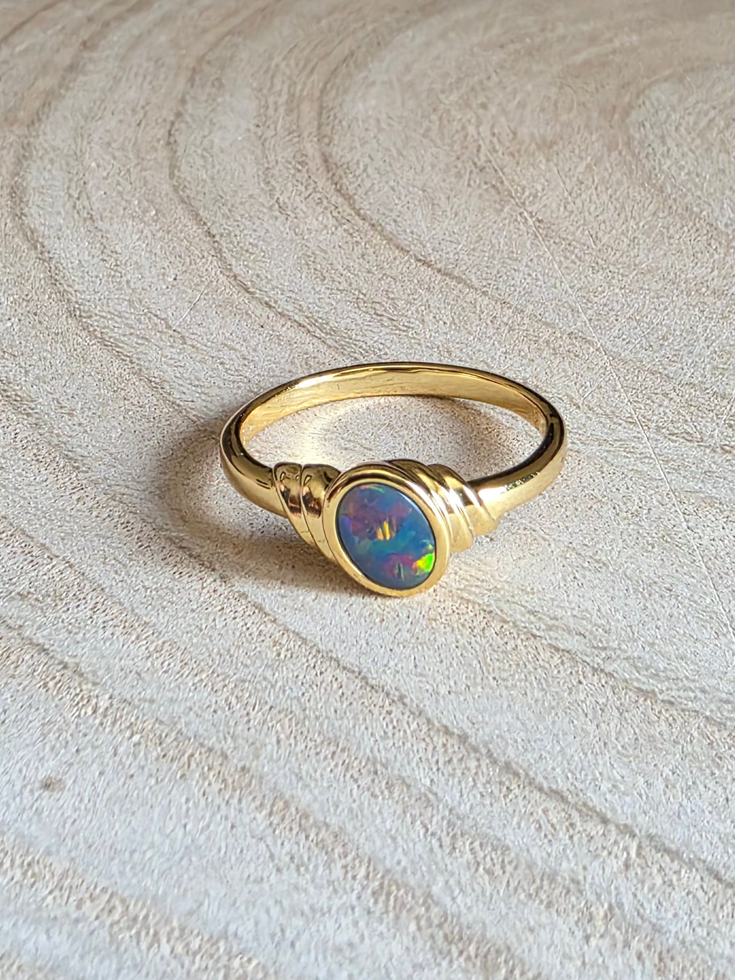 A gold ring with a round iridescent opal centre stone, resting on a light-coloured wood slice background.