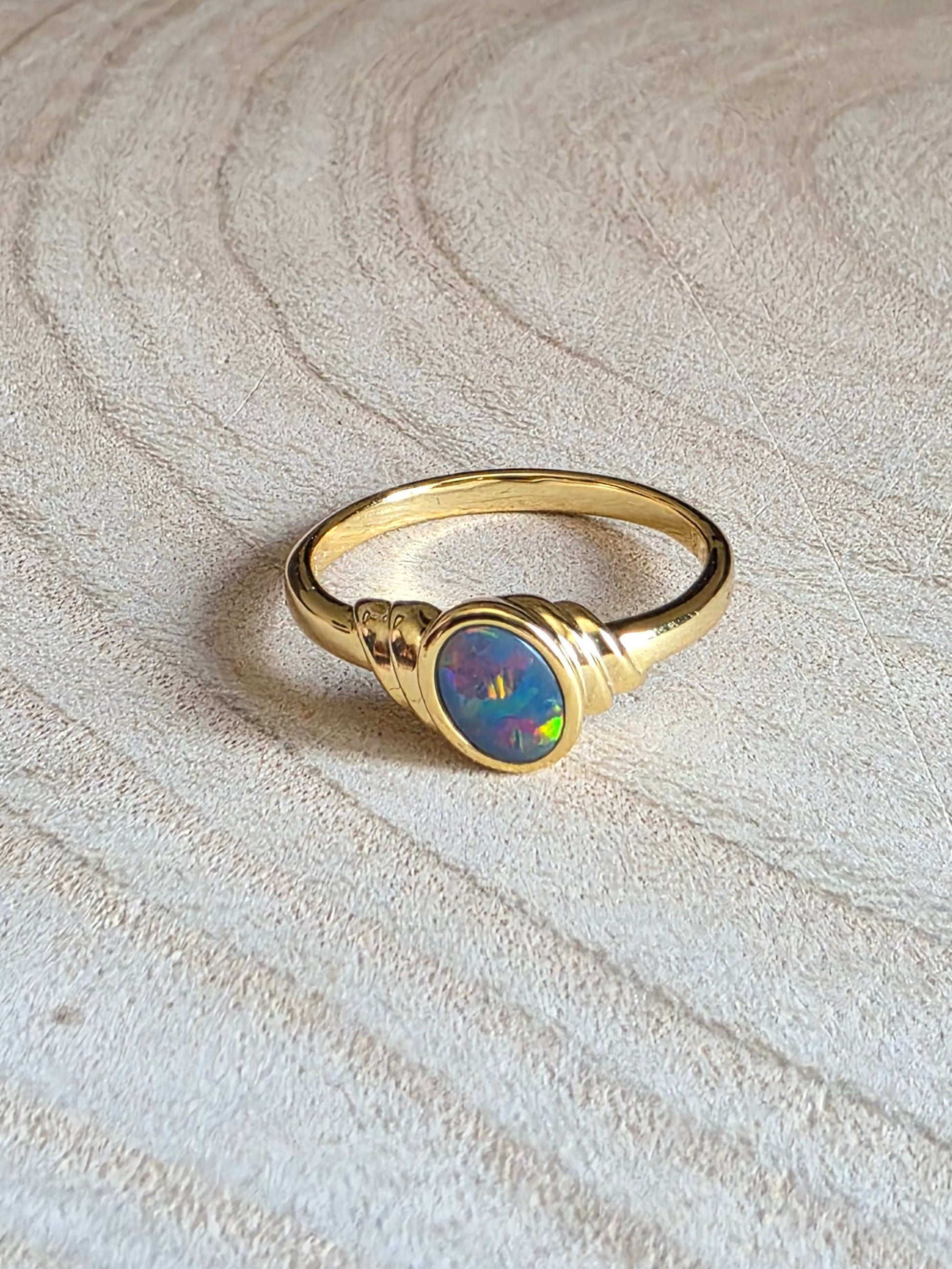 A gold ring with a round iridescent opal centre stone, resting on a light-coloured wood slice background.