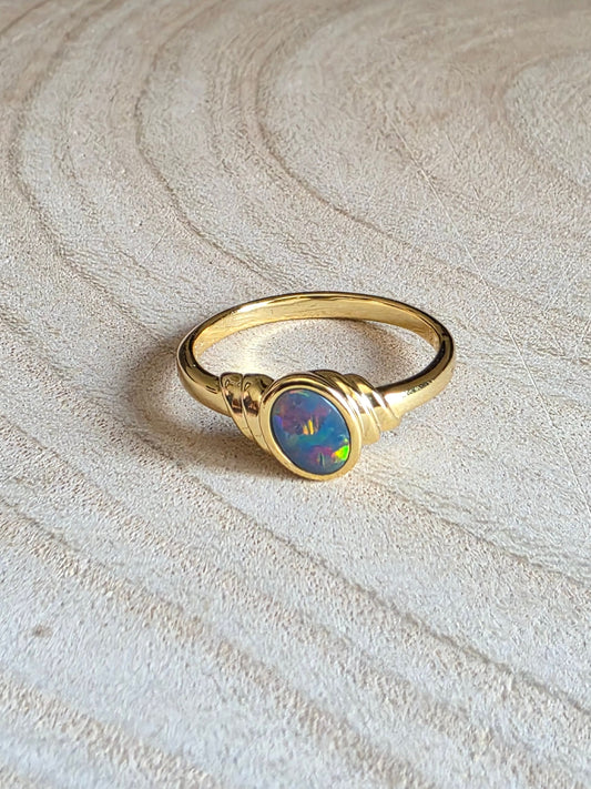 A gold ring with a round iridescent opal centre stone, resting on a light-coloured wood slice background.