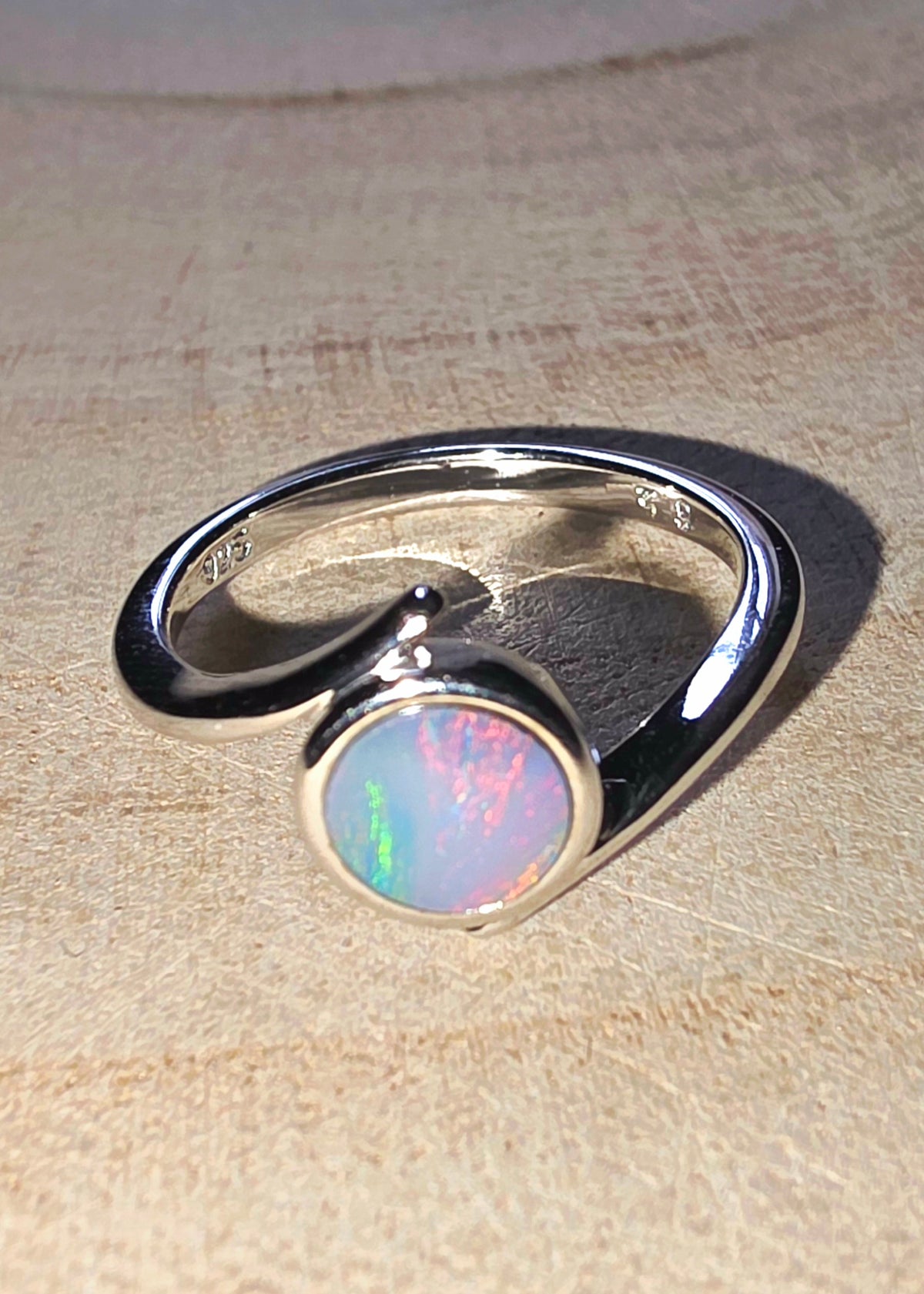 A sterling silver ring featuring a round bezel-set iridescent opal gemstone, displayed on a textured beige surface.