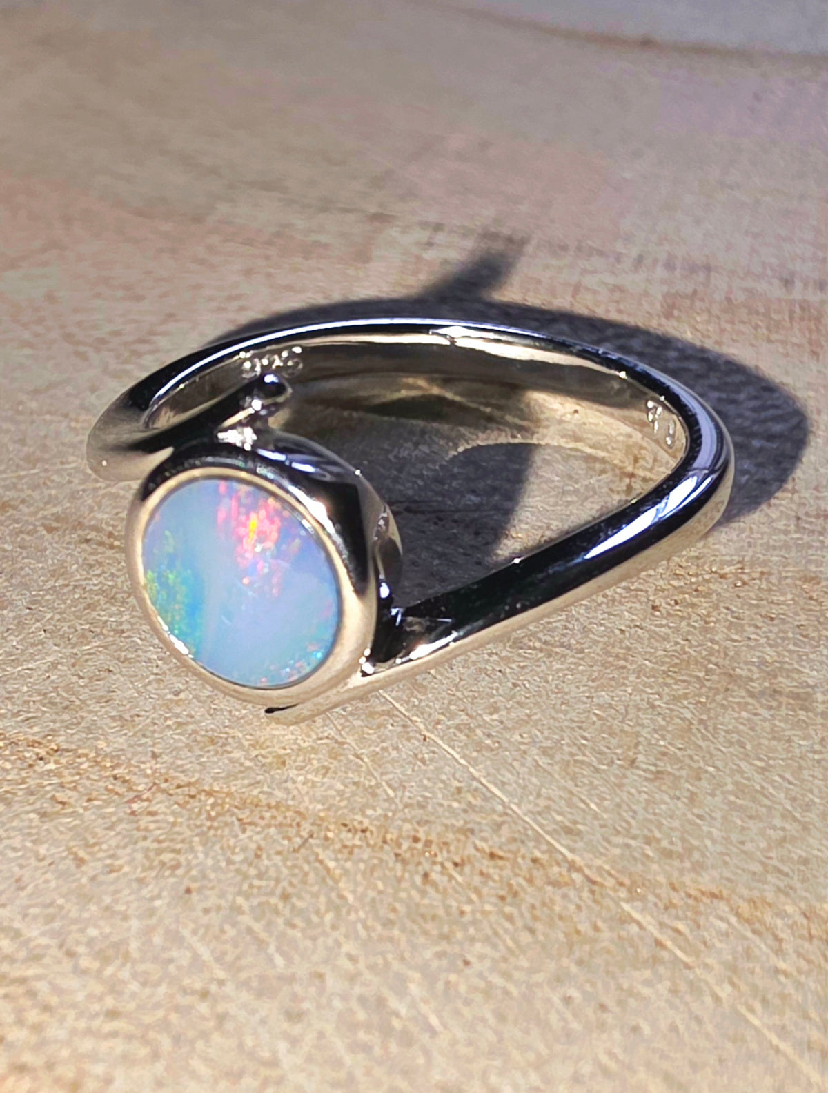 A sterling silver ring featuring a bezel-set round cabochon opal with iridescent blue, pink, and white play-of-colour, resting on a textured beige surface.
