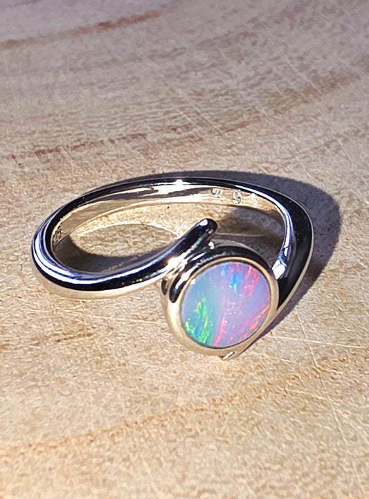 A silver open-band ring with a round iridescent opal gemstone, photographed on a light wood surface.