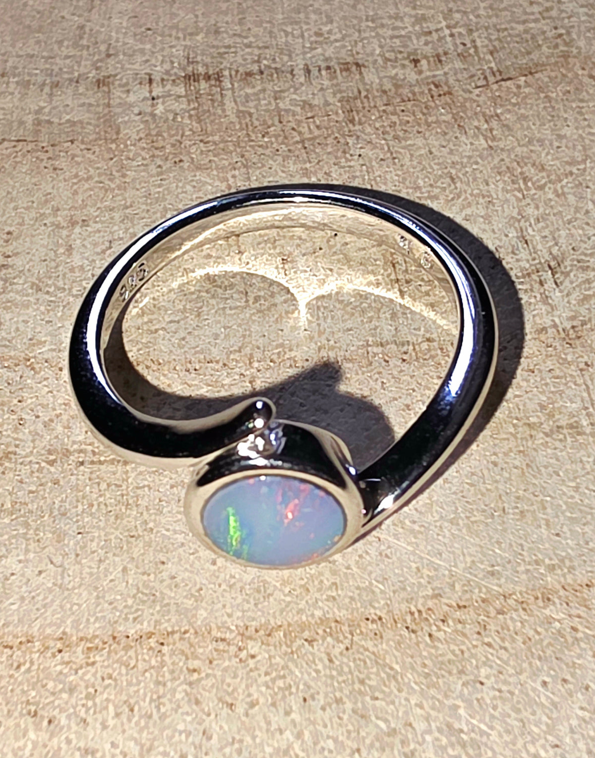 A silver-toned open ring band featuring a round cabochon opal gemstone with iridescent blue, green, and pink flashes, resting on a textured beige surface.