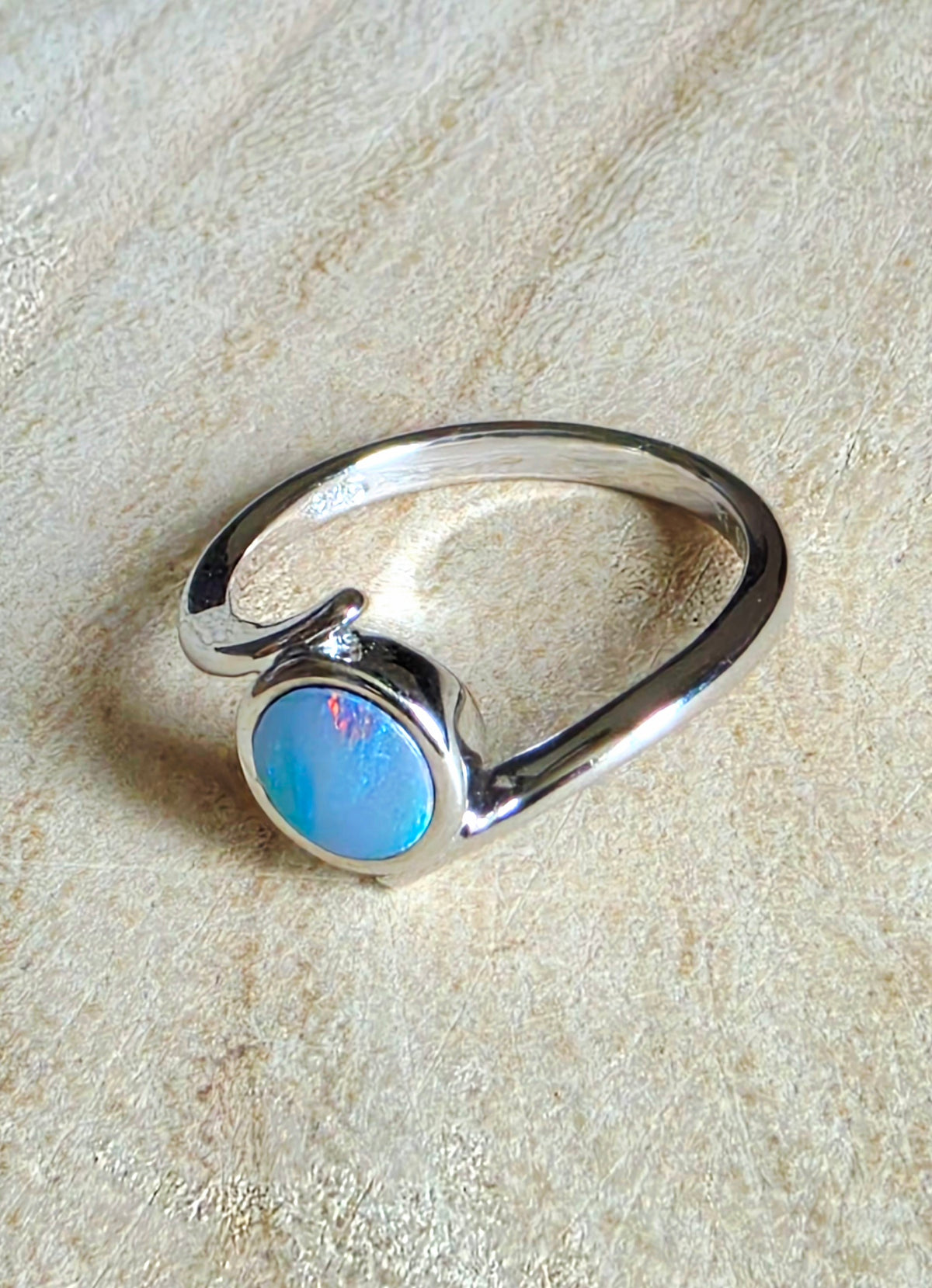 A silver ring featuring a round cabochon opal gemstone with vibrant blue, pink, and green iridescent colours, displayed on a textured beige fabric background.