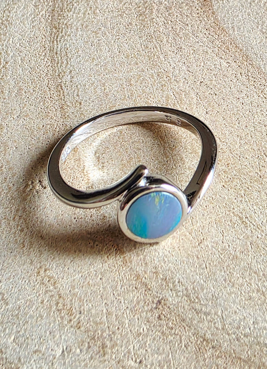 A sterling silver ring with a curved band and a round bezel-set light blue opal stone, displayed on a textured beige surface.