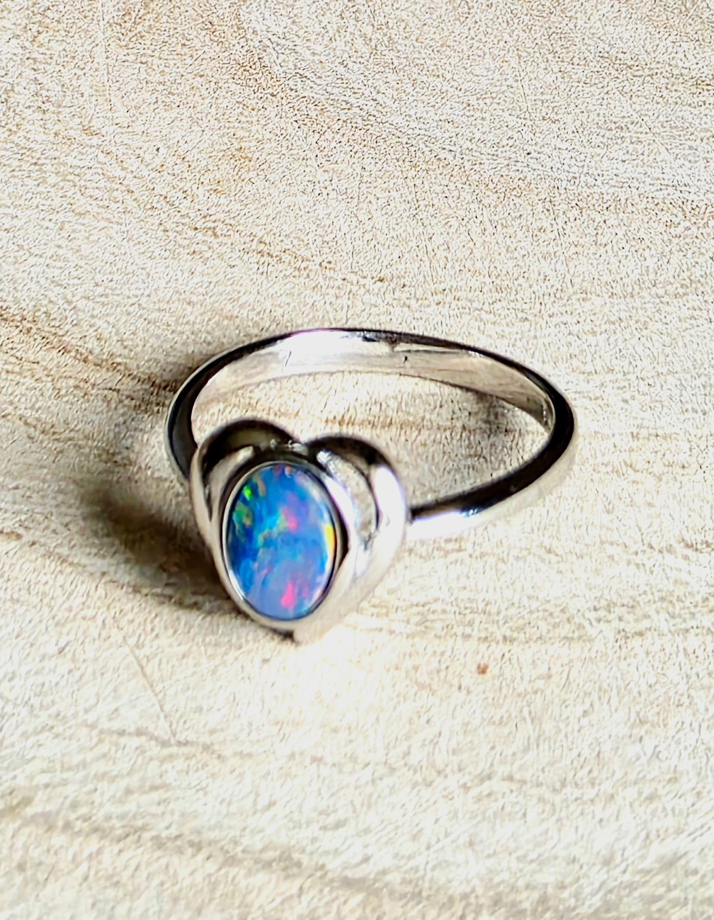 A sterling silver ring featuring a heart-shaped bezel set with a vibrant, multicoloured opal gemstone, displayed on a textured beige background.