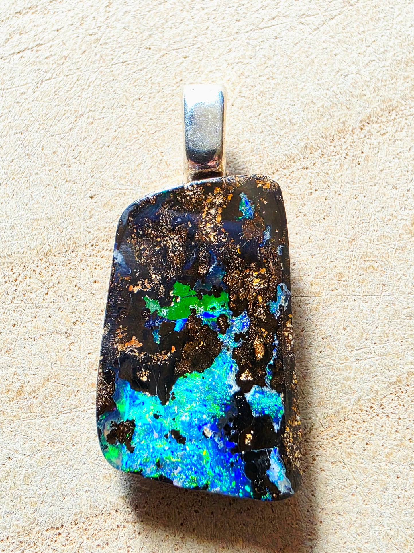 A raw, irregular-shaped opal pendant with a silver bail, showcasing vibrant blue, green, and gold flashes on a dark black base, set against a textured beige background.