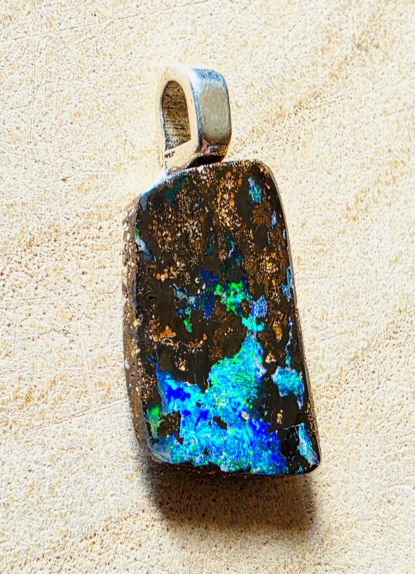A raw, rough-cut rectangular opal pendant with a silver bail, showcasing vibrant blue, green, and orange flashes on a dark brown to black base, set against a textured beige background.