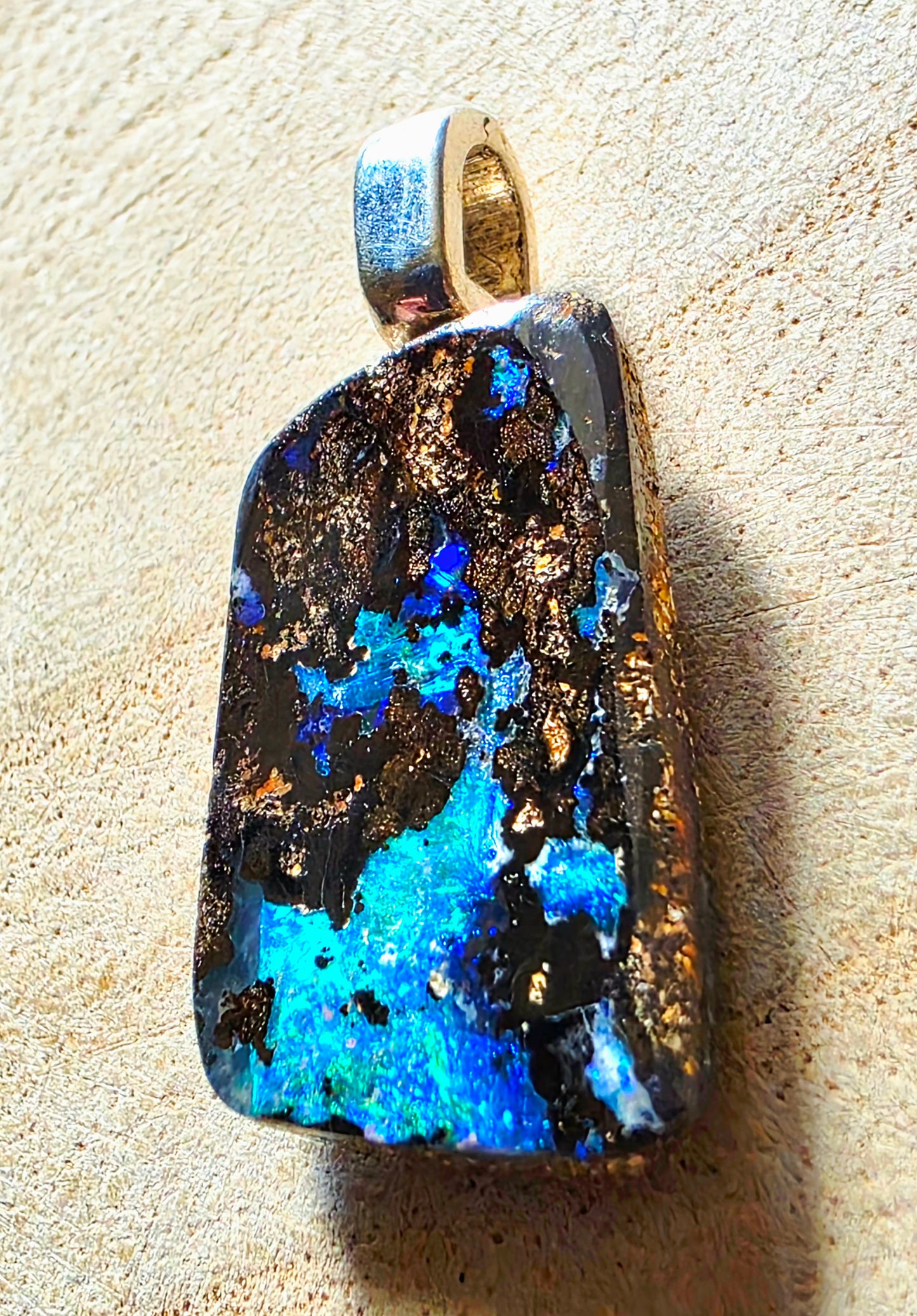 A raw opal pendant with vibrant blue and turquoise flashes, dark black matrix, and gold flakes, set in a silver bail, resting on a textured beige surface.