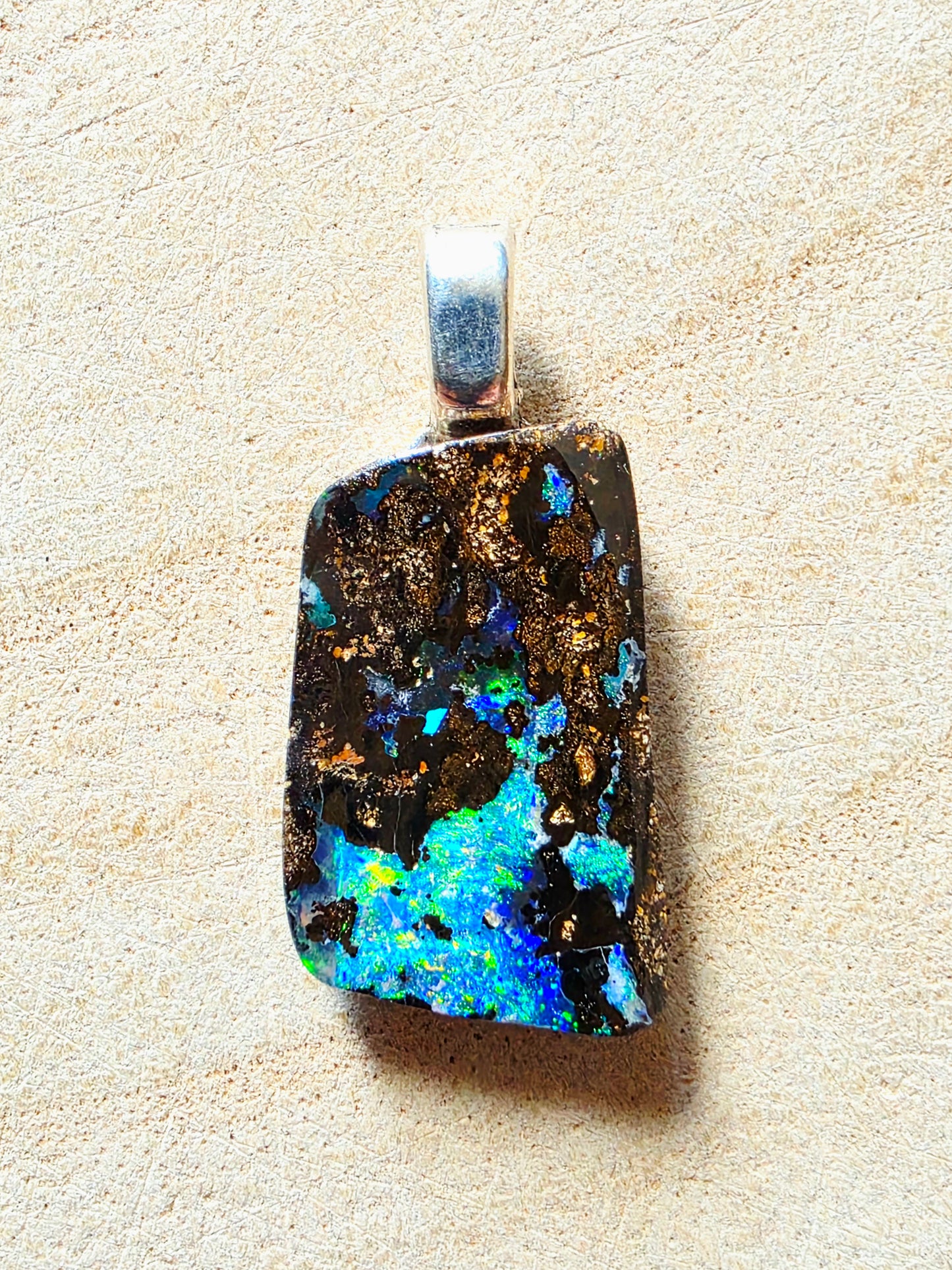 A rectangular silver pendant featuring a highly iridescent, opal-like stone with vibrant blue, green, and yellow flashes over a dark brown and black matrix with gold flecks, set against a light beige textured background.