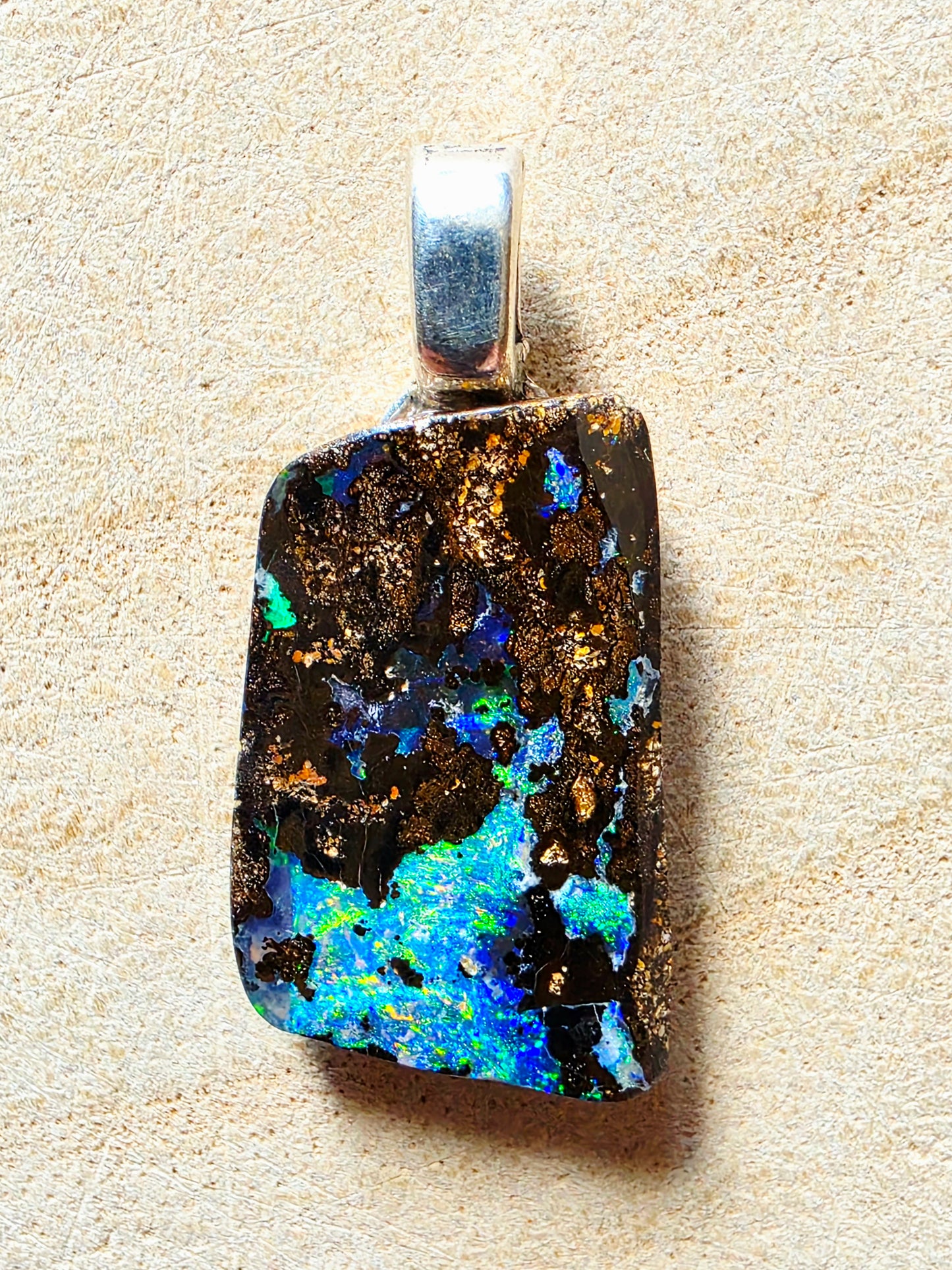 A rectangular silver pendant featuring a natural black opal stone with vibrant blue, green, and gold flashes, displayed on a textured beige background.
