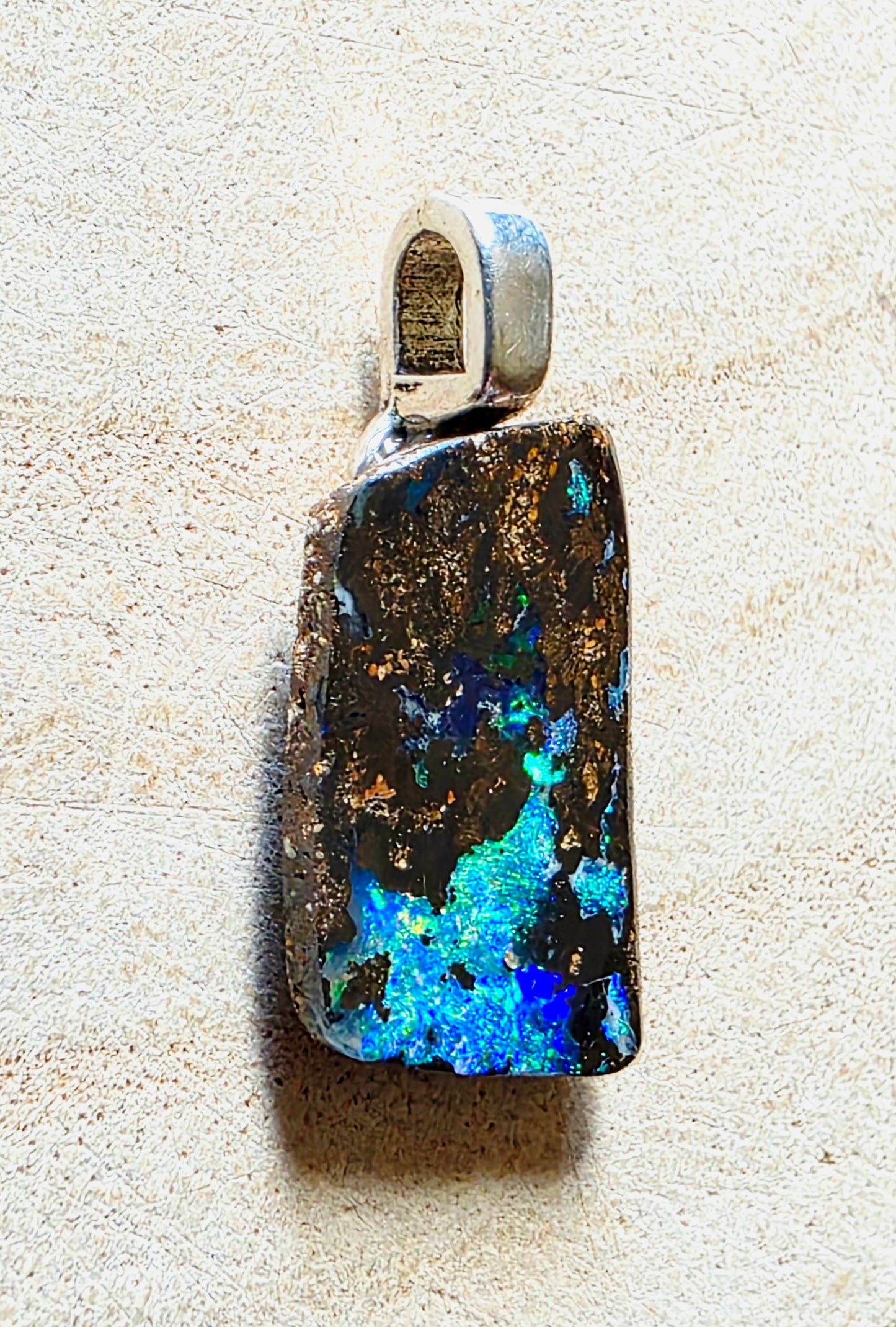 A rectangular opal pendant with a silver bail, resting on a textured beige surface. The opal displays vibrant blue, green, and black play-of-colour with golden flecks.