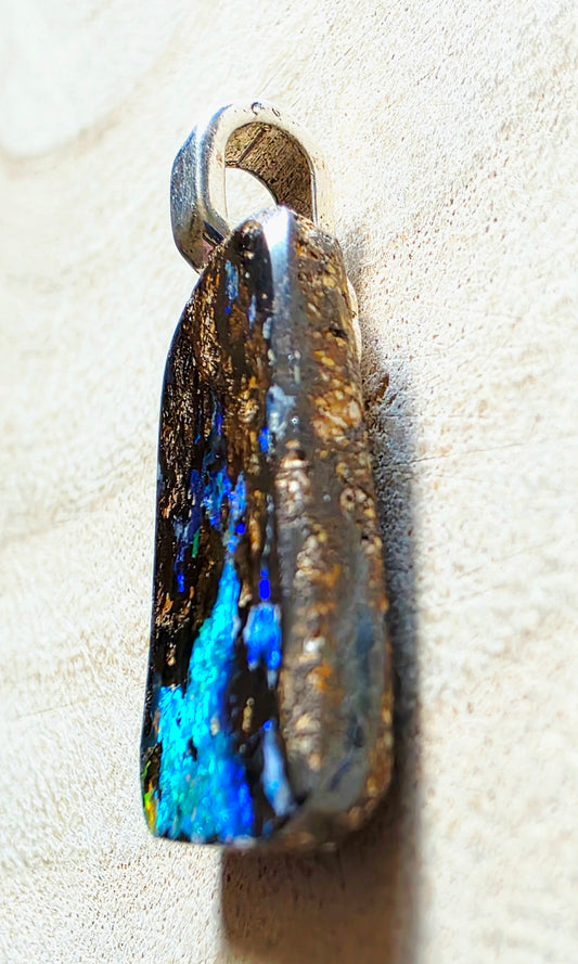 A raw opal pendant with a silver bail, resting on a textured beige surface. The pendant features a natural, rough-cut opal stone showing vibrant blue, green, and gold iridescence against a dark base, with a polished silver metal loop at the top.