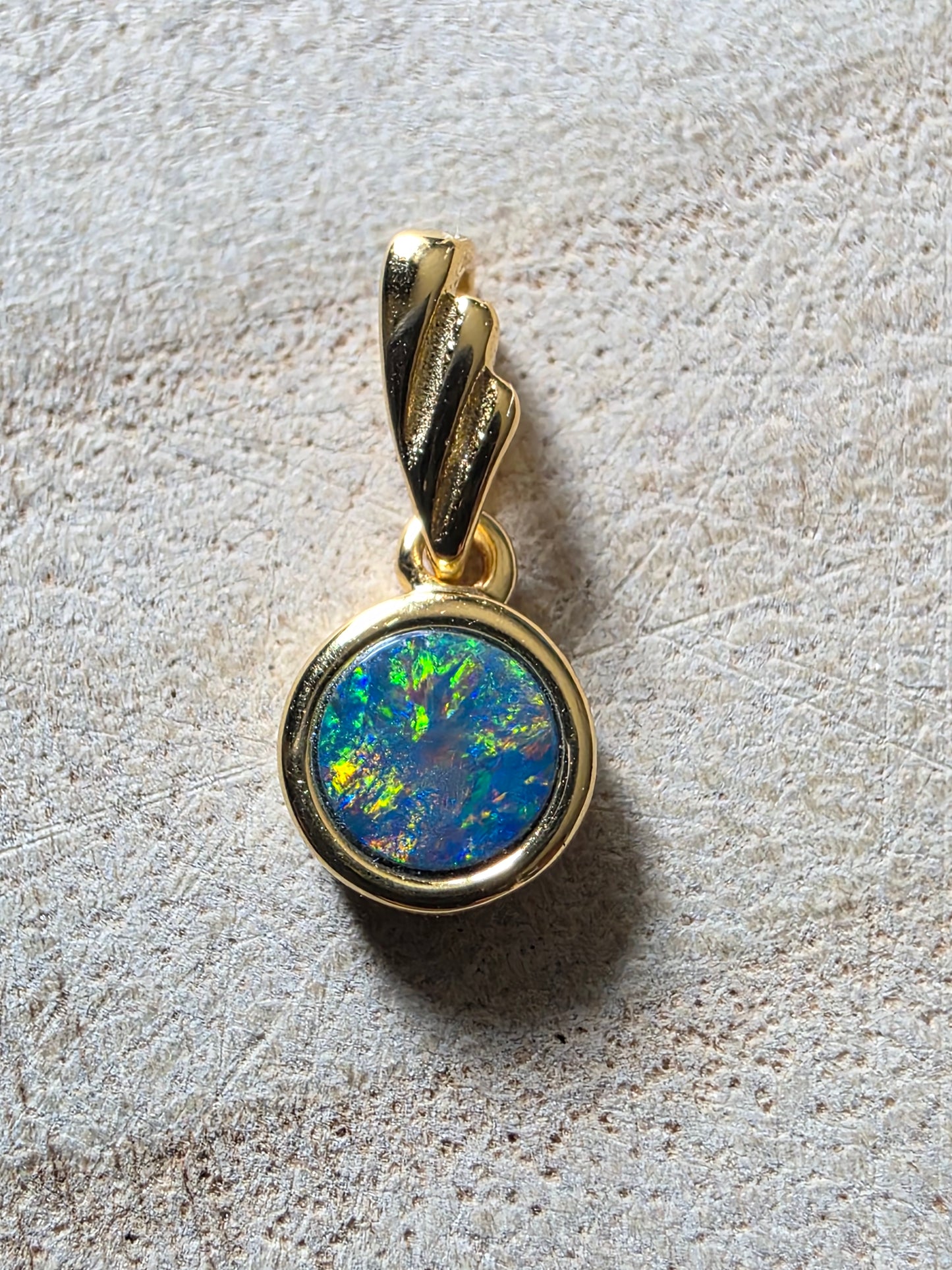 A gold pendant featuring a round cabochon opal with vibrant blue, green, and orange flashes, set on a textured light beige surface.