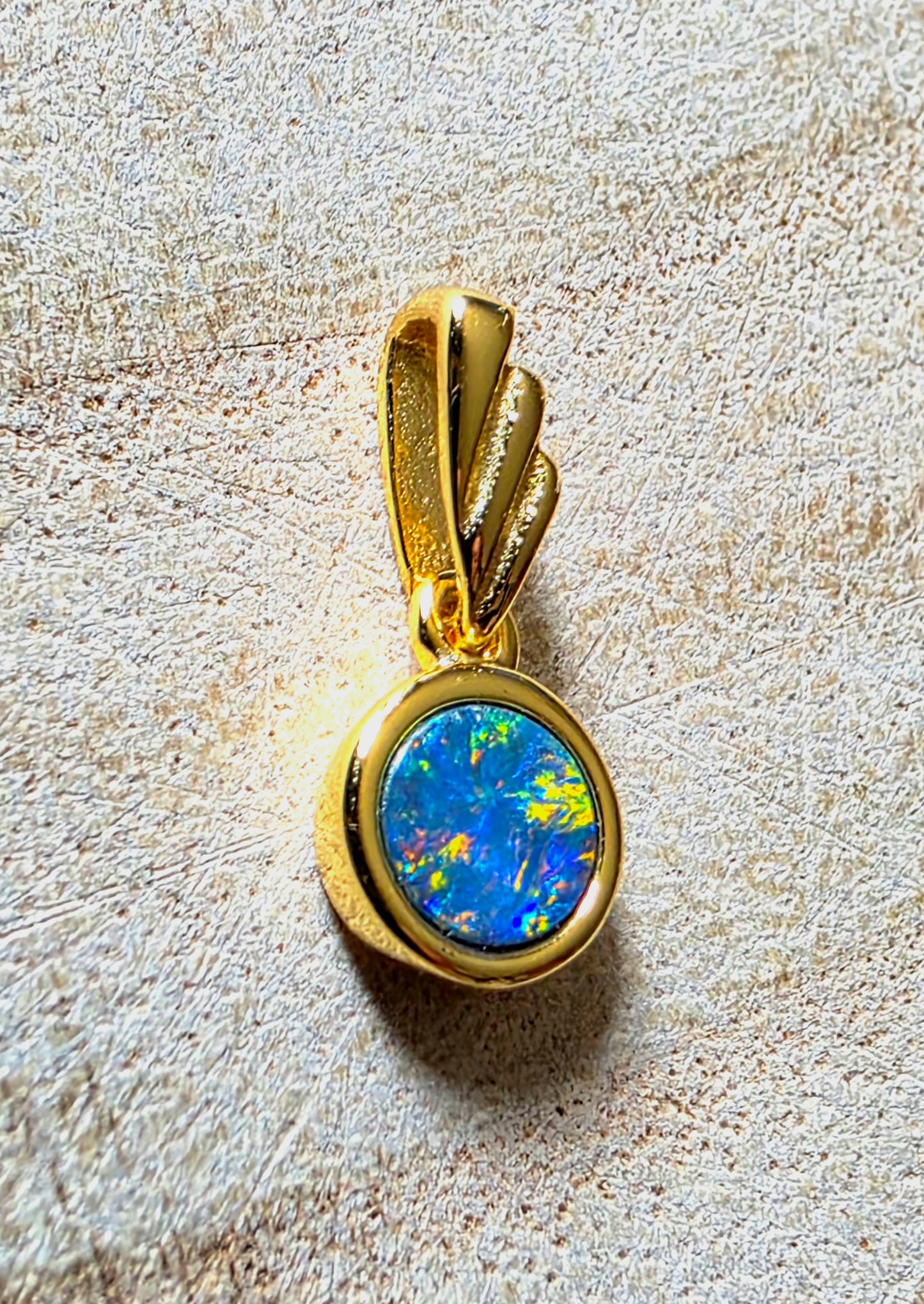 A gold pendant featuring a round blue opal stone with vibrant green and yellow flashes, set in a polished gold bezel on a textured beige background.