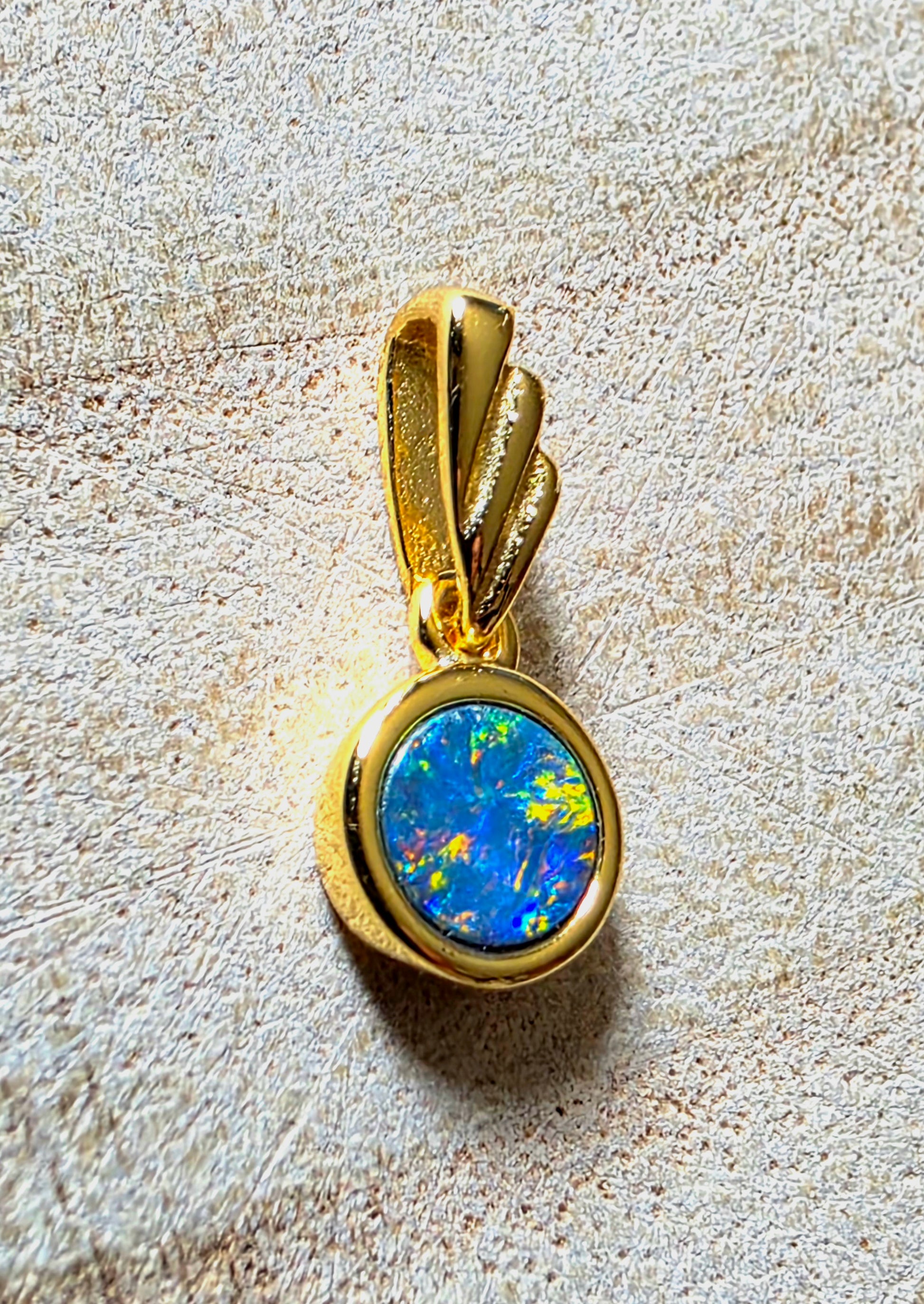 A gold pendant featuring a round blue opal stone with vibrant green and yellow flashes, set in a polished gold bezel on a textured beige background.