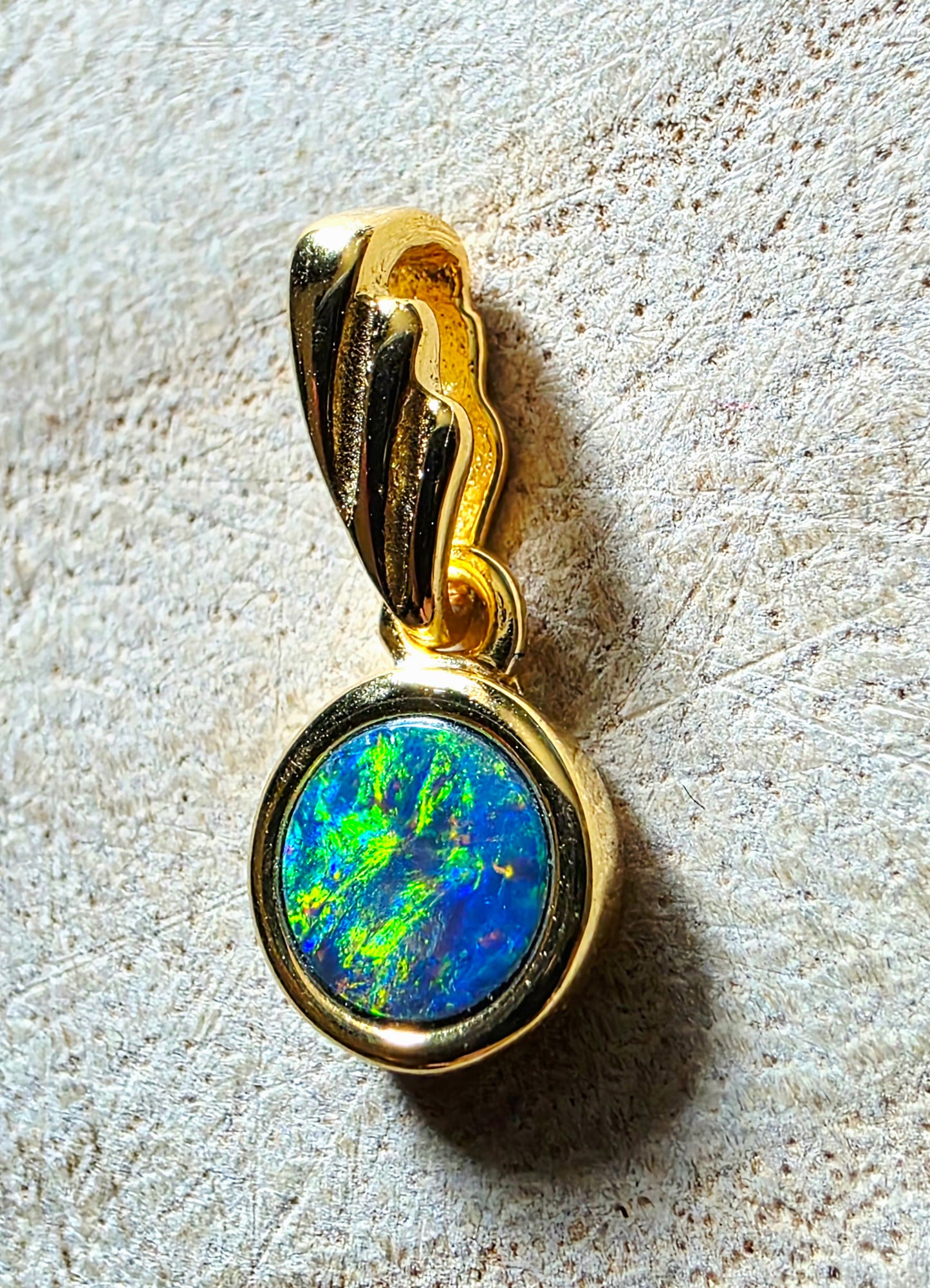 A gold pendant featuring a round bezel-set iridescent opal with vibrant blue, green, and orange flashes, resting on a textured beige surface.