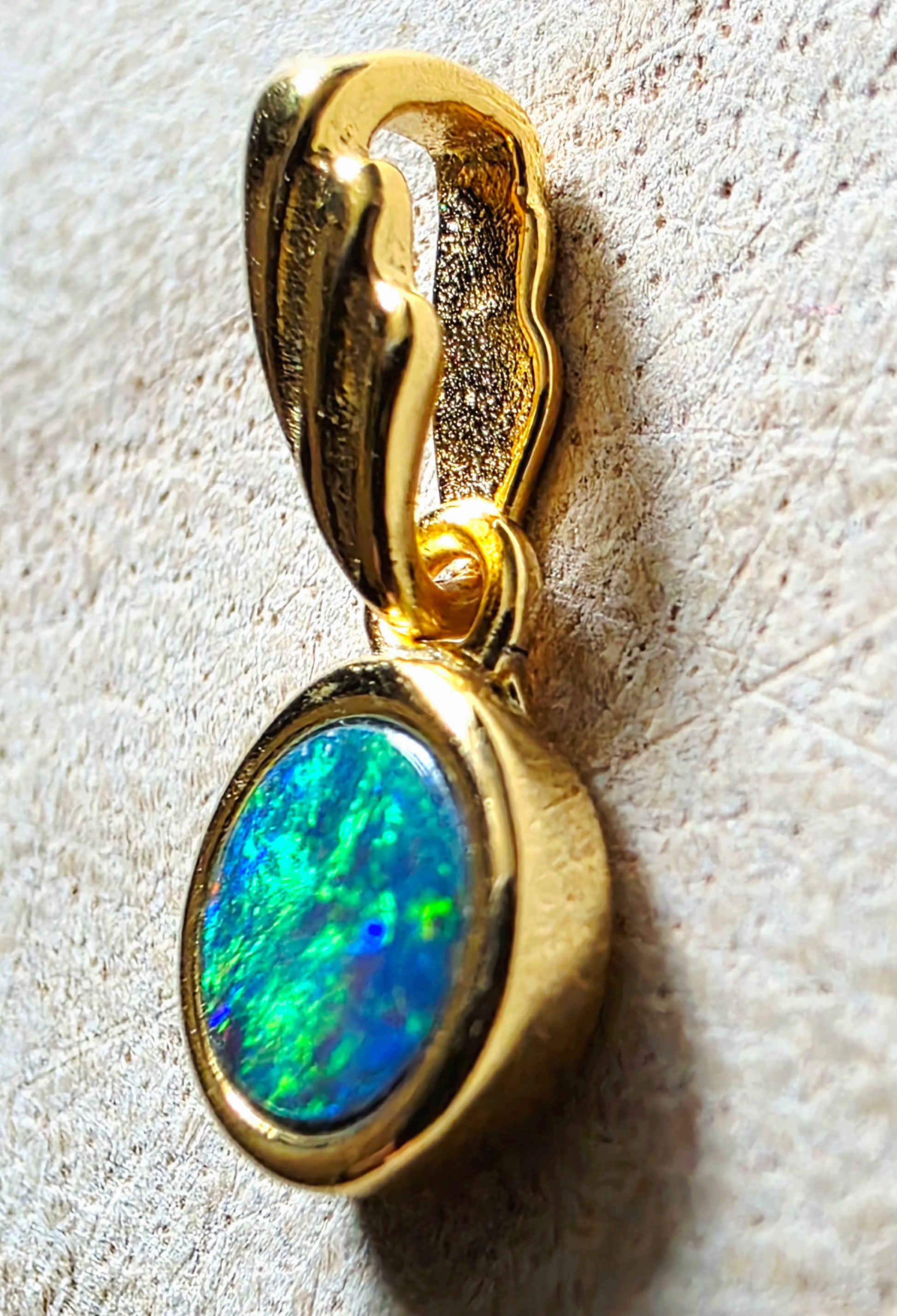 A gold pendant featuring an iridescent opal stone with vibrant blue, green, and orange flashes, set in a polished gold bezel with a decorative wavy bail above it, resting on a textured light beige surface.