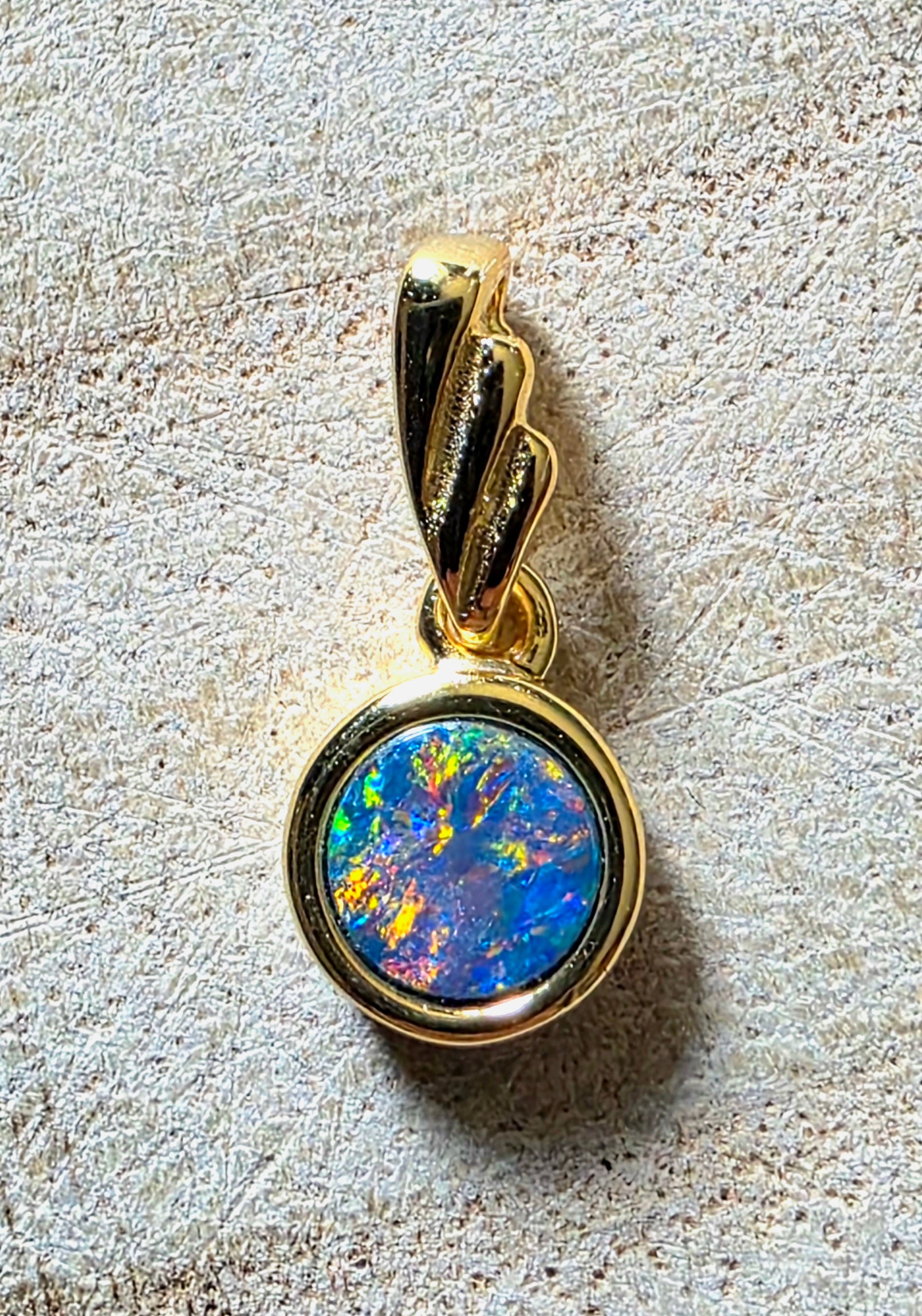 A gold pendant featuring a round bezel-set iridescent opal with vibrant blue, green, orange, and yellow flashes, shown on a textured light beige stone background.