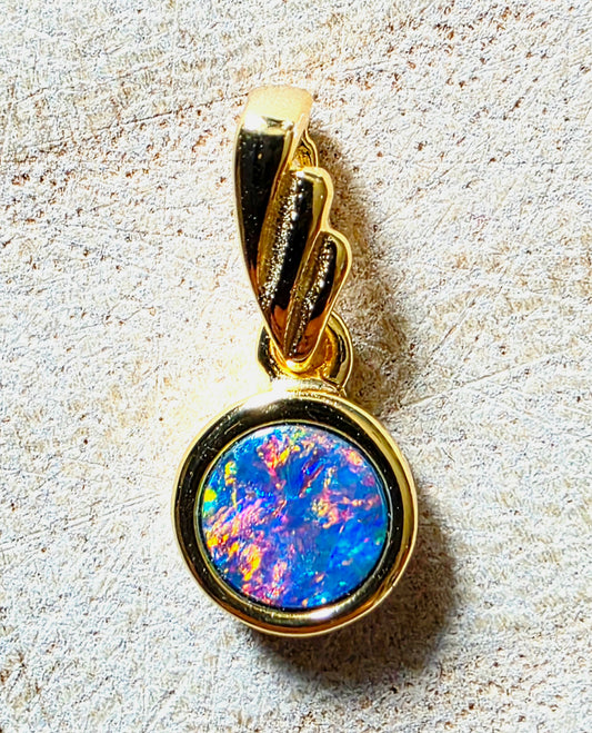 A gold pendant featuring a round iridescent opal stone with vibrant blue, orange, and purple flashes, set in a polished gold bezel with a twisted bail above.