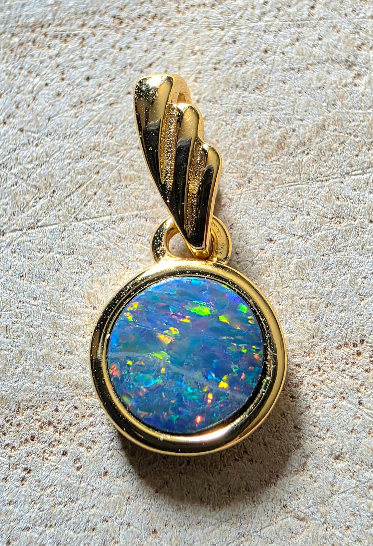 A gold pendant featuring a round, vibrant blue and green opal gemstone, photographed on a textured beige surface.