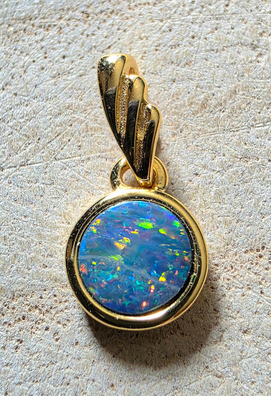 A gold pendant featuring a round, vibrant blue and green opal gemstone, photographed on a textured beige surface.