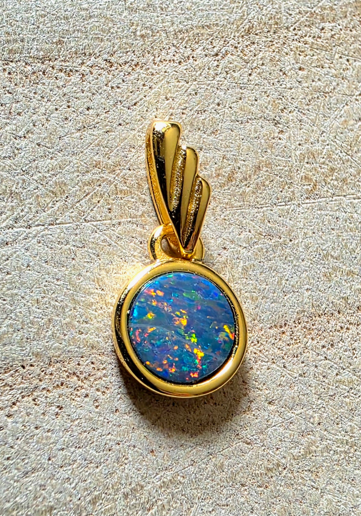 A gold pendant featuring a round cabochon opal gemstone with vibrant blue, green, and orange play-of-colour, set in a polished gold bezel with a gold bail, displayed on a textured beige surface.