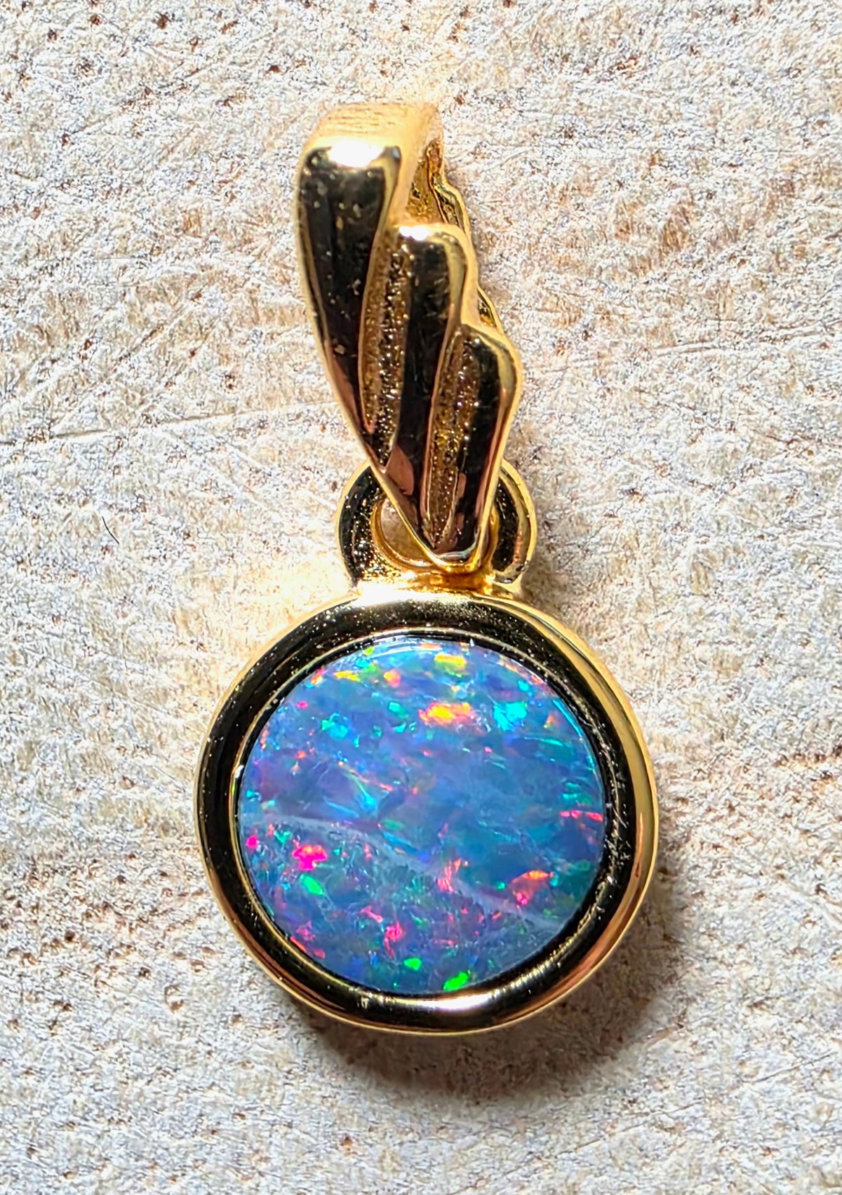 A gold pendant featuring a round bezel-set iridescent opal stone with vibrant blue, pink, green, and orange flashes, shown on a textured light beige surface.