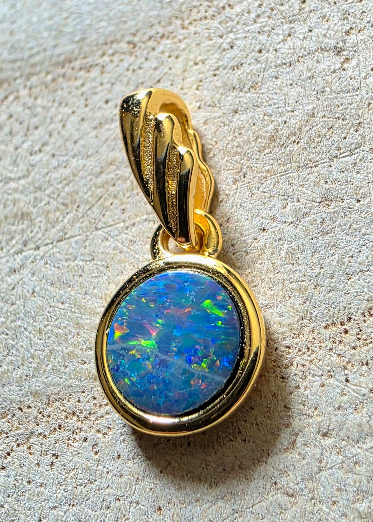 A gold pendant featuring a round, vibrant blue opal gemstone with multicoloured flashes, set in a polished gold bezel with a textured gold bail, resting on a light beige textured surface.
