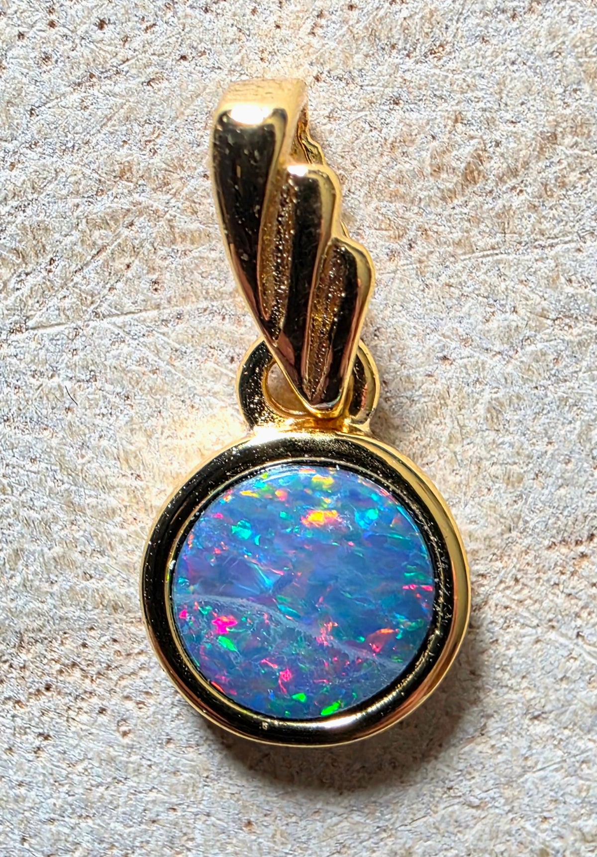 A gold pendant featuring a round cabochon opal with vibrant blue, green, pink, and orange flashes, set in a polished gold bezel with a decorative gold bail, displayed on a textured beige surface.