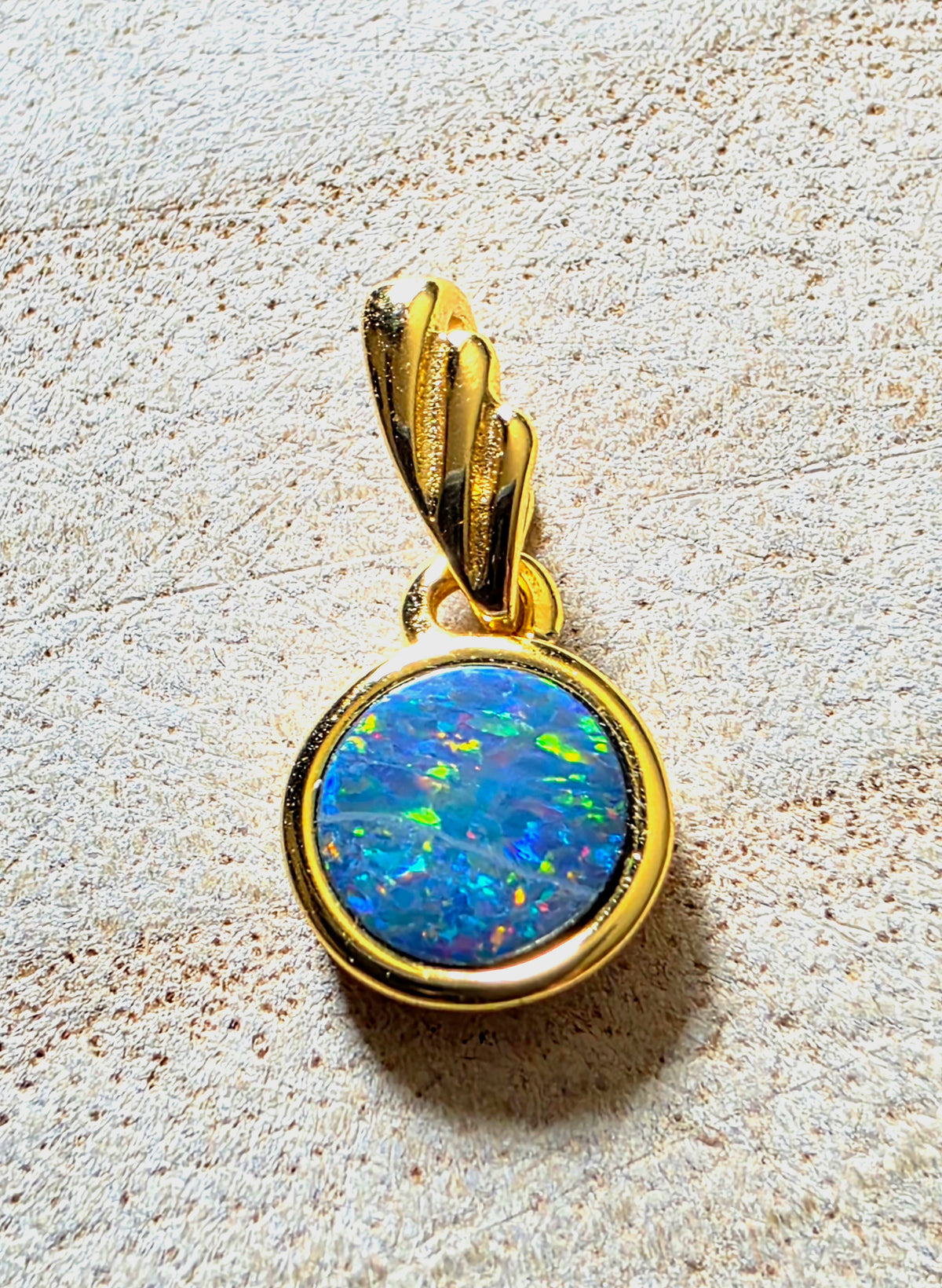 A gold pendant featuring a round, vibrant blue opal gemstone in a gold bezel setting, suspended from a textured gold bail, displayed on a textured beige stone surface.