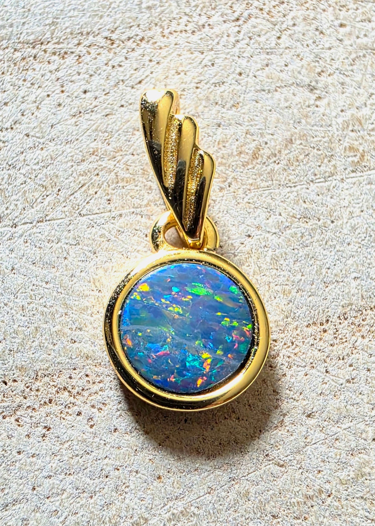 A gold pendant featuring a round, vibrant blue opal cabochon with multicoloured flashes, set in a polished gold bezel with a textured gold bail, displayed on a light beige textured surface.