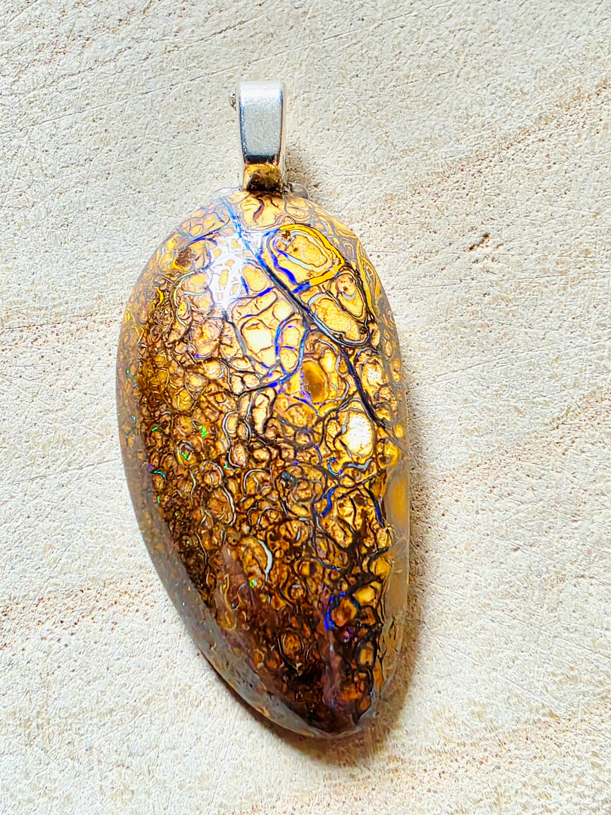 An oval pendant with a highly iridescent, mottled pattern of gold, brown, and blue, set in a silver-toned metal bail and resting on a textured beige surface.