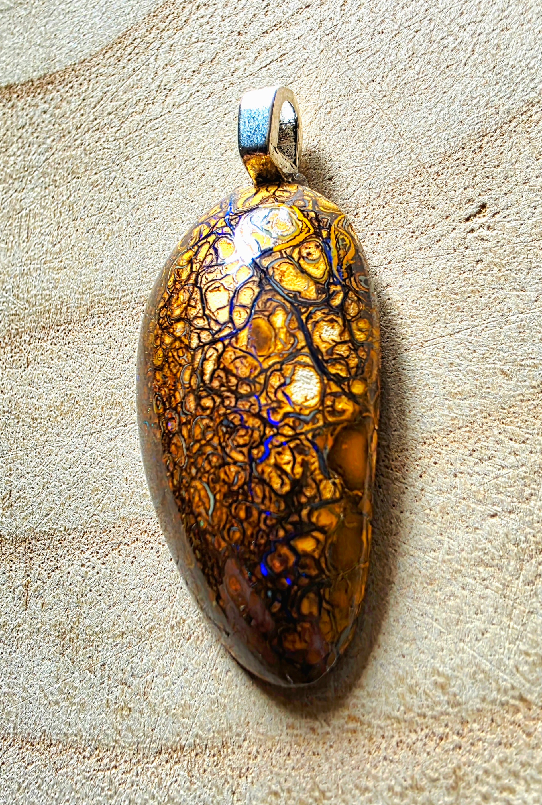 A teardrop-shaped pendant featuring a highly iridescent, golden-orange body with a dark branching network pattern and subtle blue flashes, suspended from a silver bail, set against a textured beige background.