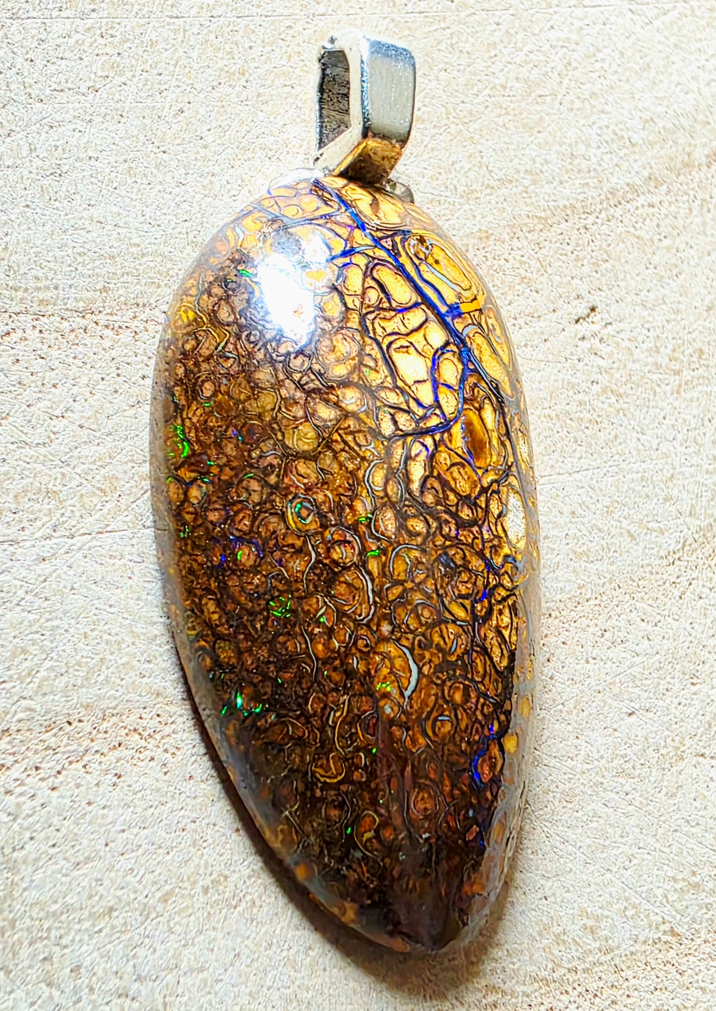 A teardrop-shaped pendant featuring a highly iridescent, patterned opal-like stone with gold, brown, and blue network patterns, suspended from a silver bail, set against a textured beige background.