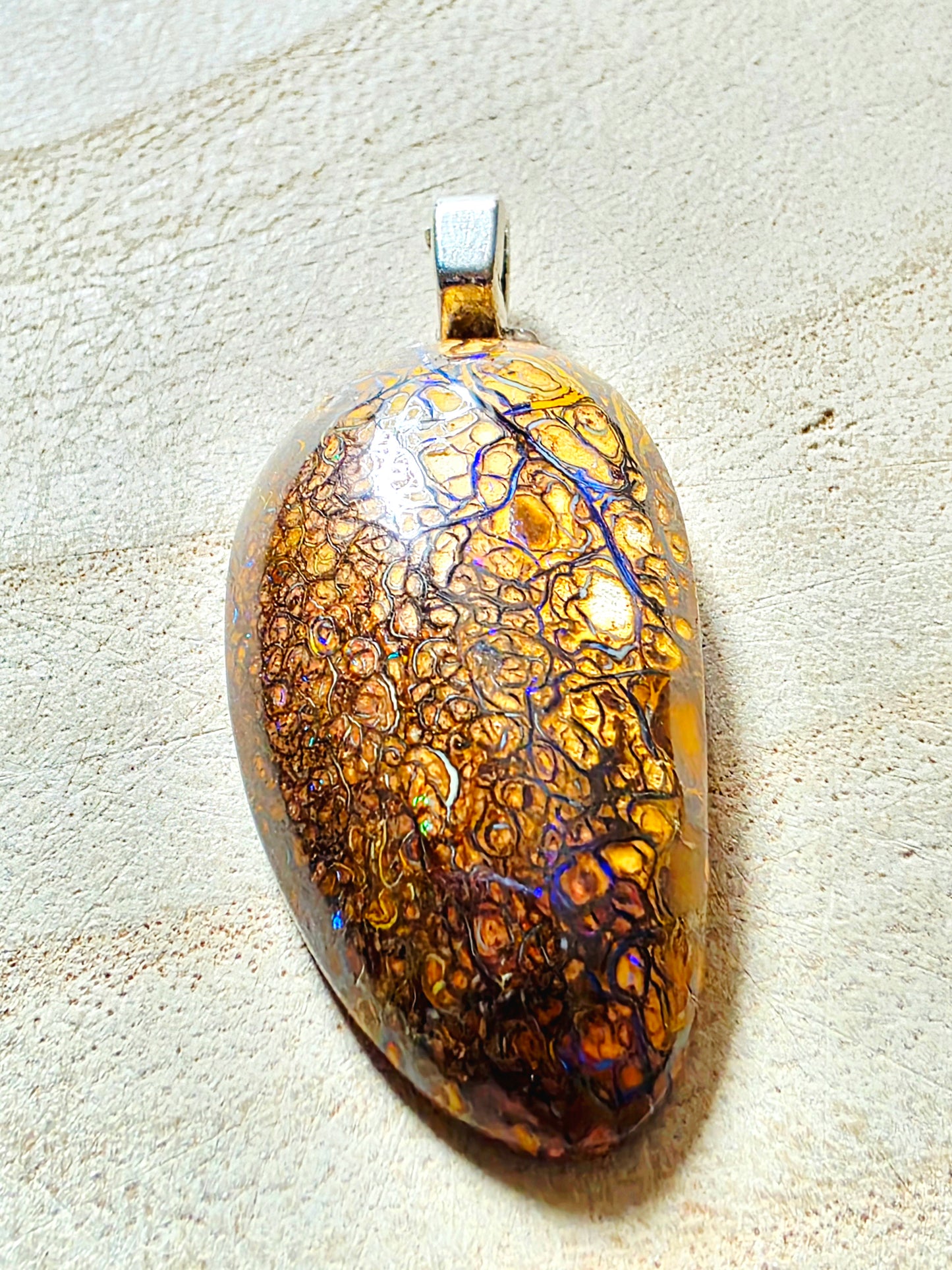 A pendant featuring a polished brown and amber bohemian opal stone with intricate web-like patterns and blue iridescent veins, suspended from a silver bail, set against a textured beige background.