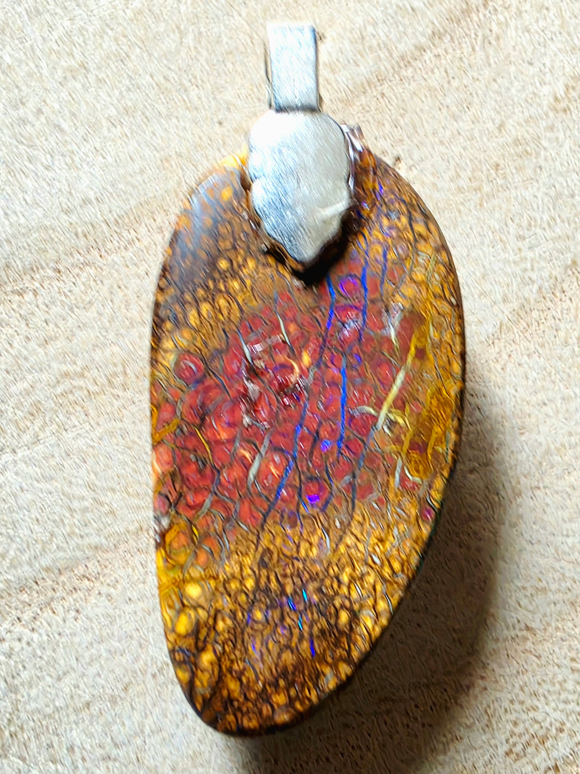 A irregularly shaped pendant featuring a mottled pattern of gold, brown, red, and iridescent blue-orange flashes, suspended from a silver-coloured metal cap on a textured beige background.