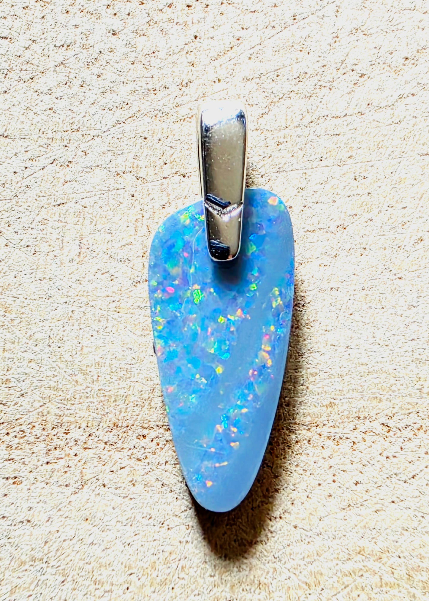 A light blue opal pendant with iridescent flashes and a silver bail, shown on a textured beige background.