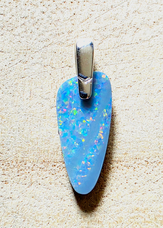A light blue opal pendant with iridescent flashes and a silver bail, shown on a textured beige background.