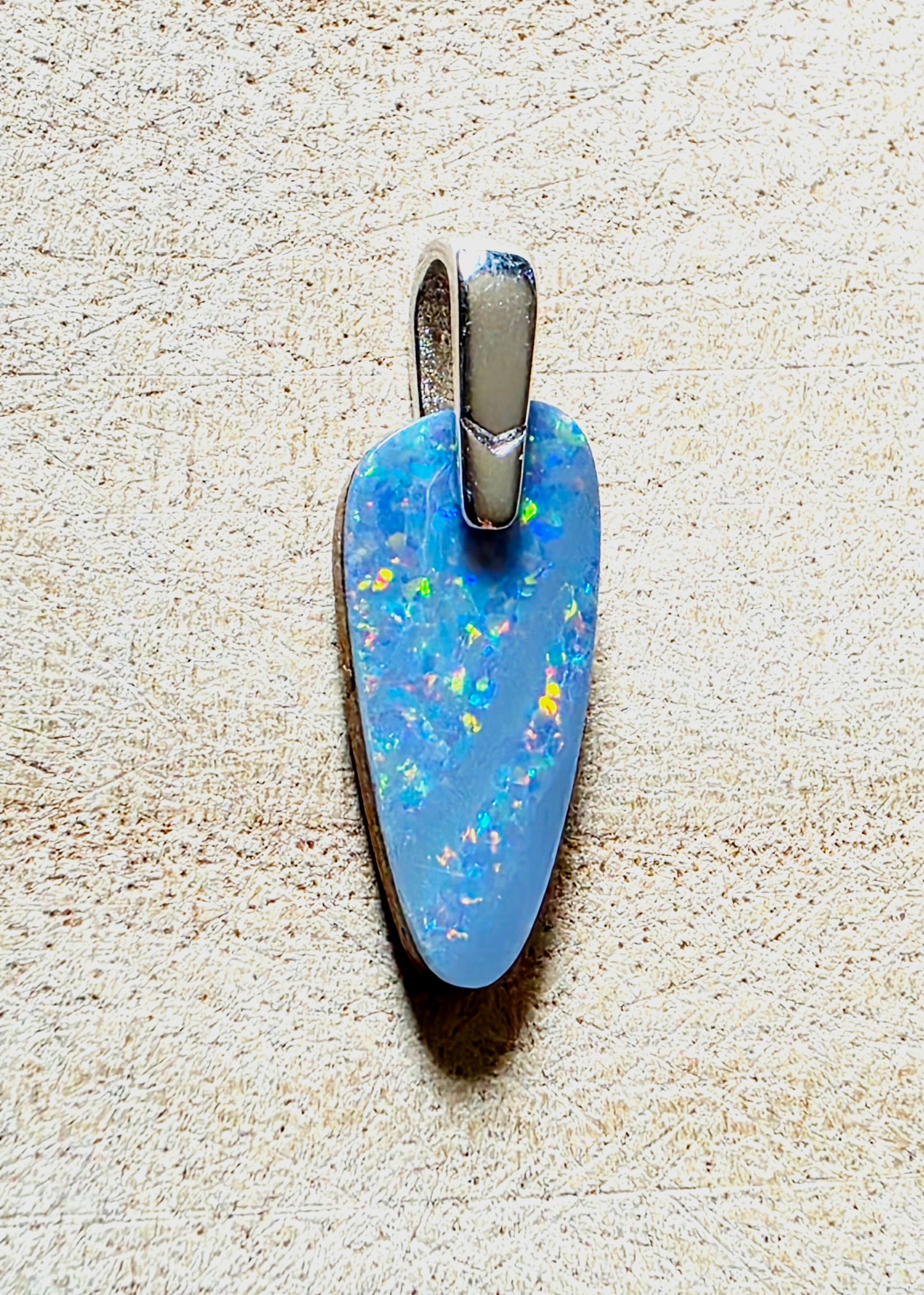 A teardrop-shaped blue opal pendant with a silver bail, resting on a textured beige surface.
